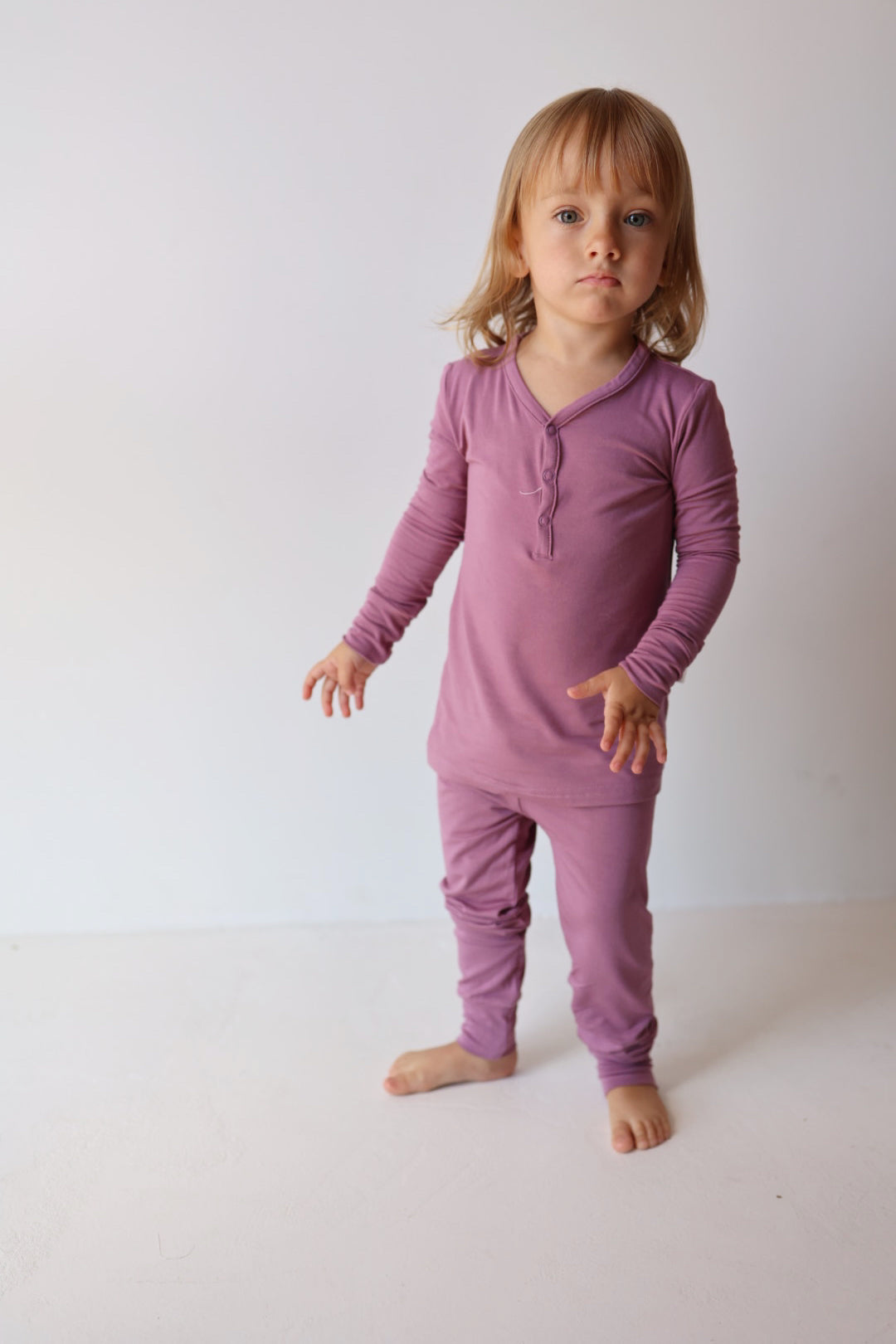 A young child with shoulder-length blond hair stands barefoot on a plain white floor against a light-colored wall. The child is wearing the Bamboo Two Piece Pajamas in Amethyst from forever french baby and looks directly at the camera with a neutral expression.