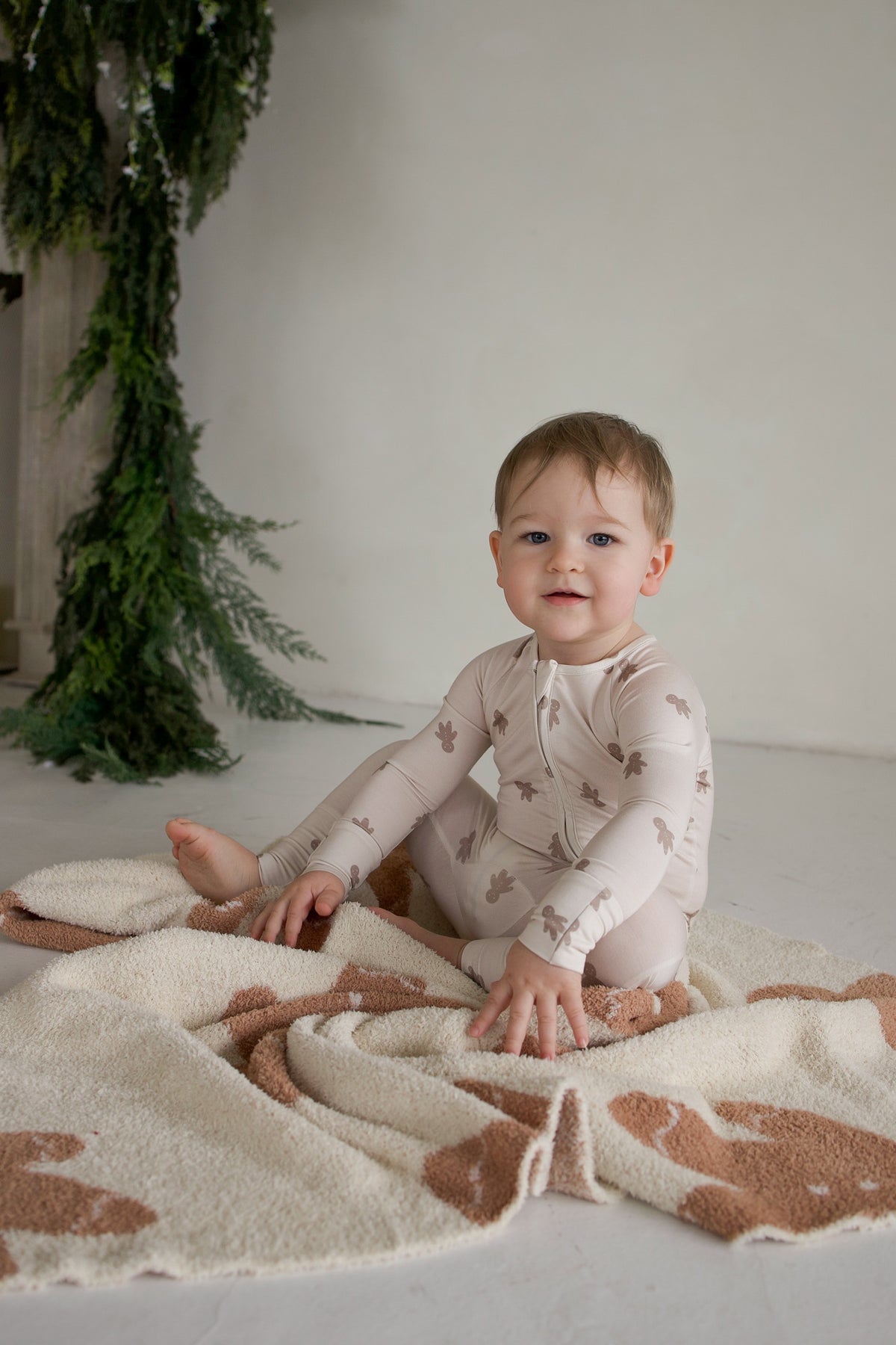 A toddler wearing Bamboo Zip Pajamas | Gingersnap with a brown gingerbread man print sits on a plush patterned blanket.