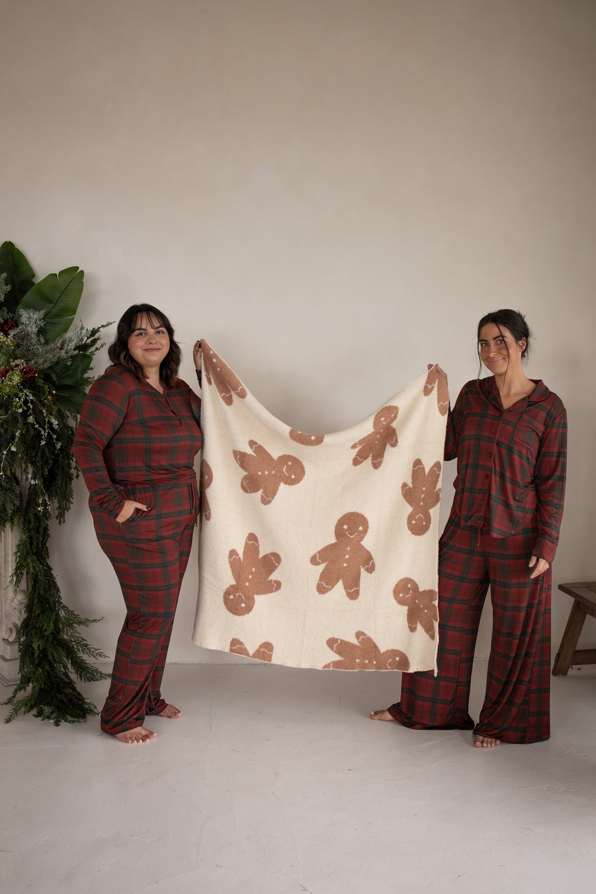 Two women in matching red plaid pajamas hold up a soft, Oeko-Tex certified *PRE-ORDER* Plush Blanket | Gingersnap patterned with brown gingerbread men.