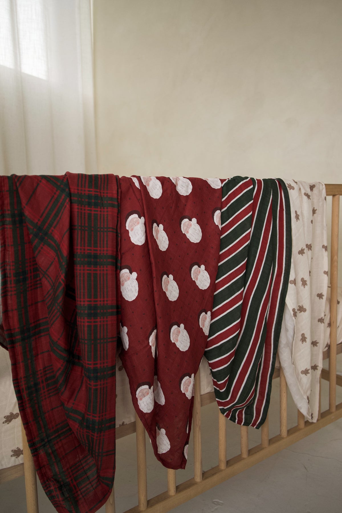 Four ultra-soft Muslin Swaddle | Christmas Plaid cotton swaddles featuring holiday patterns—red and green plaid, Santa Claus faces, stripes, and gingerbread men—draped over a wooden crib rail.
