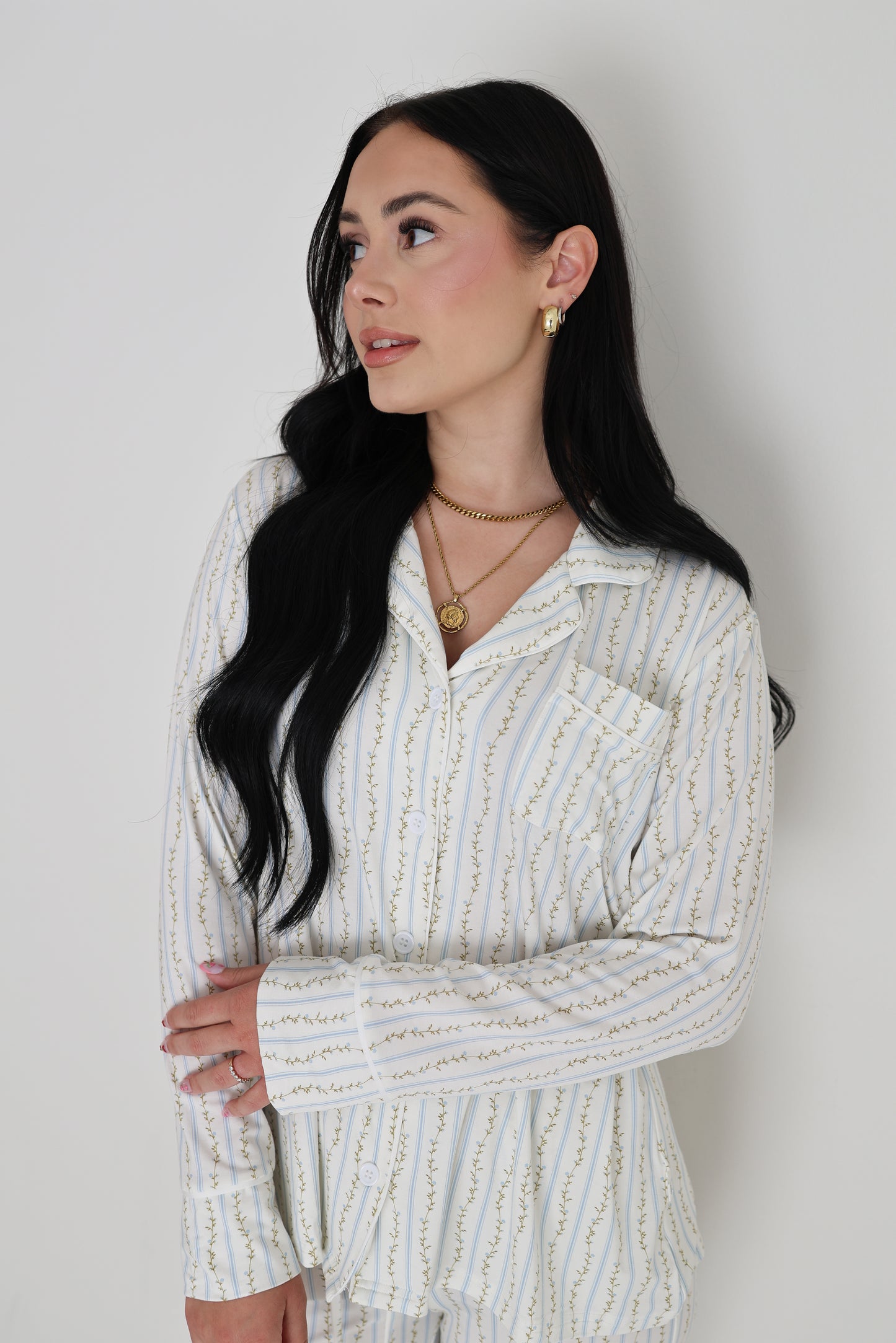Woman with long black hair wearing the forever french baby Women's Bamboo Button Up Lounge Set in Dainty Vines, featuring striped pajamas with a pocket, layered gold necklaces, and gold earrings, standing against a plain white background.