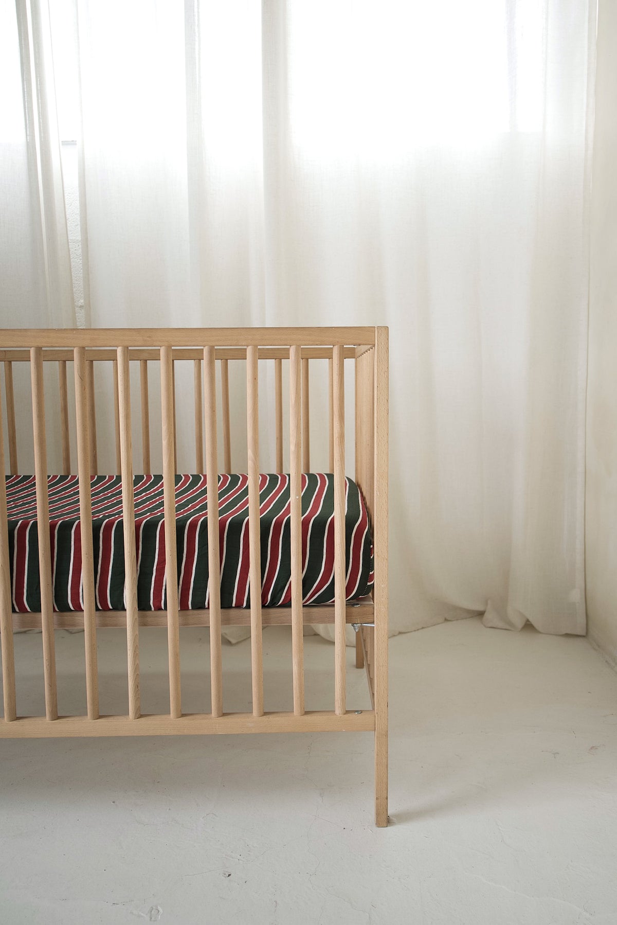 A light-wood crib featuring a Muslin Crib Sheet | Holiday Stripe with thin red, white, and black stripes.