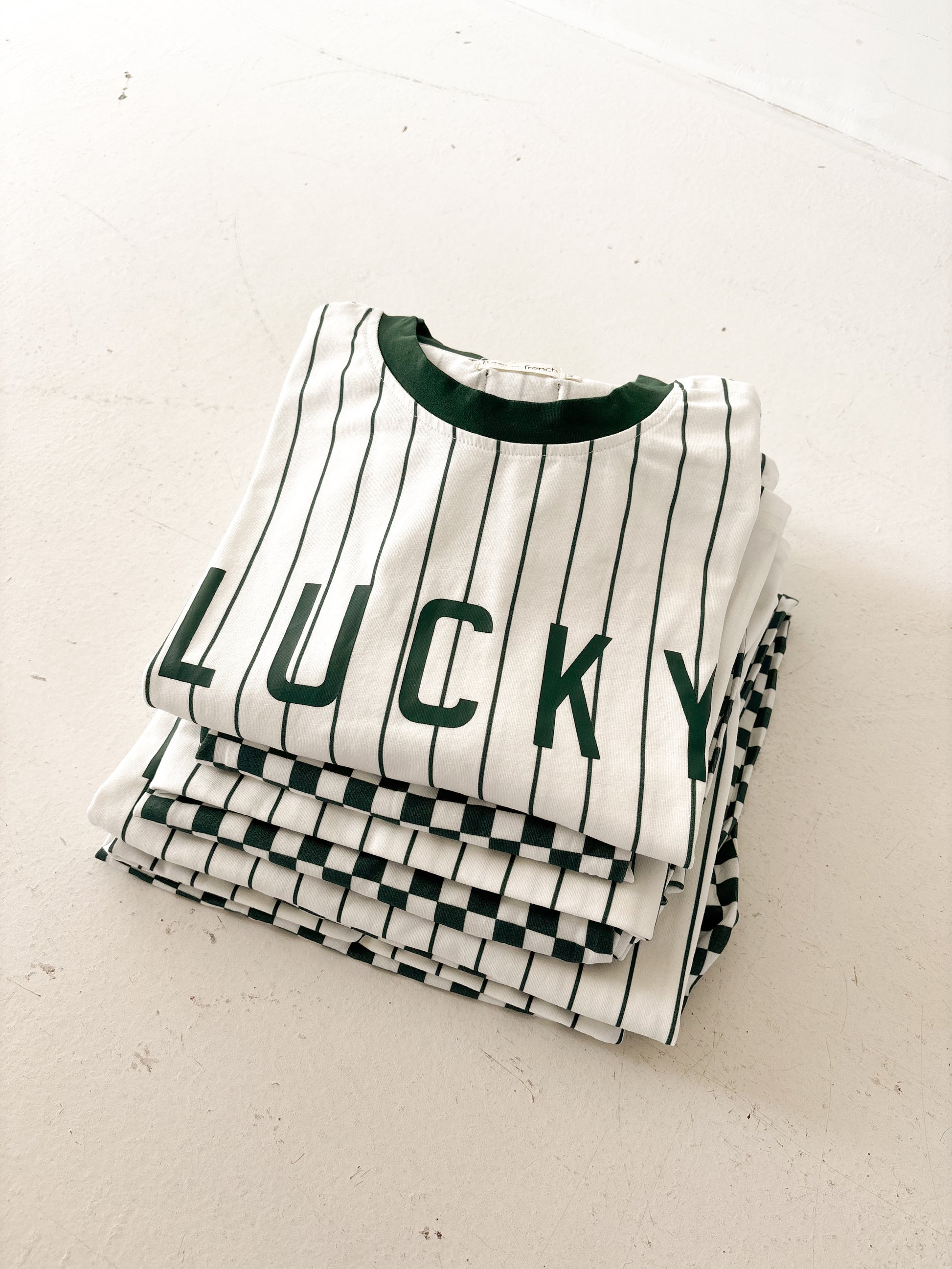 The forever french baby T-Shirt & Pant Set | Lucky Micro Check features a stack of white tees with black stripes, dark green collars, and “LUCKY” in bold green letters—perfectly paired with kids’ wide leg pants.