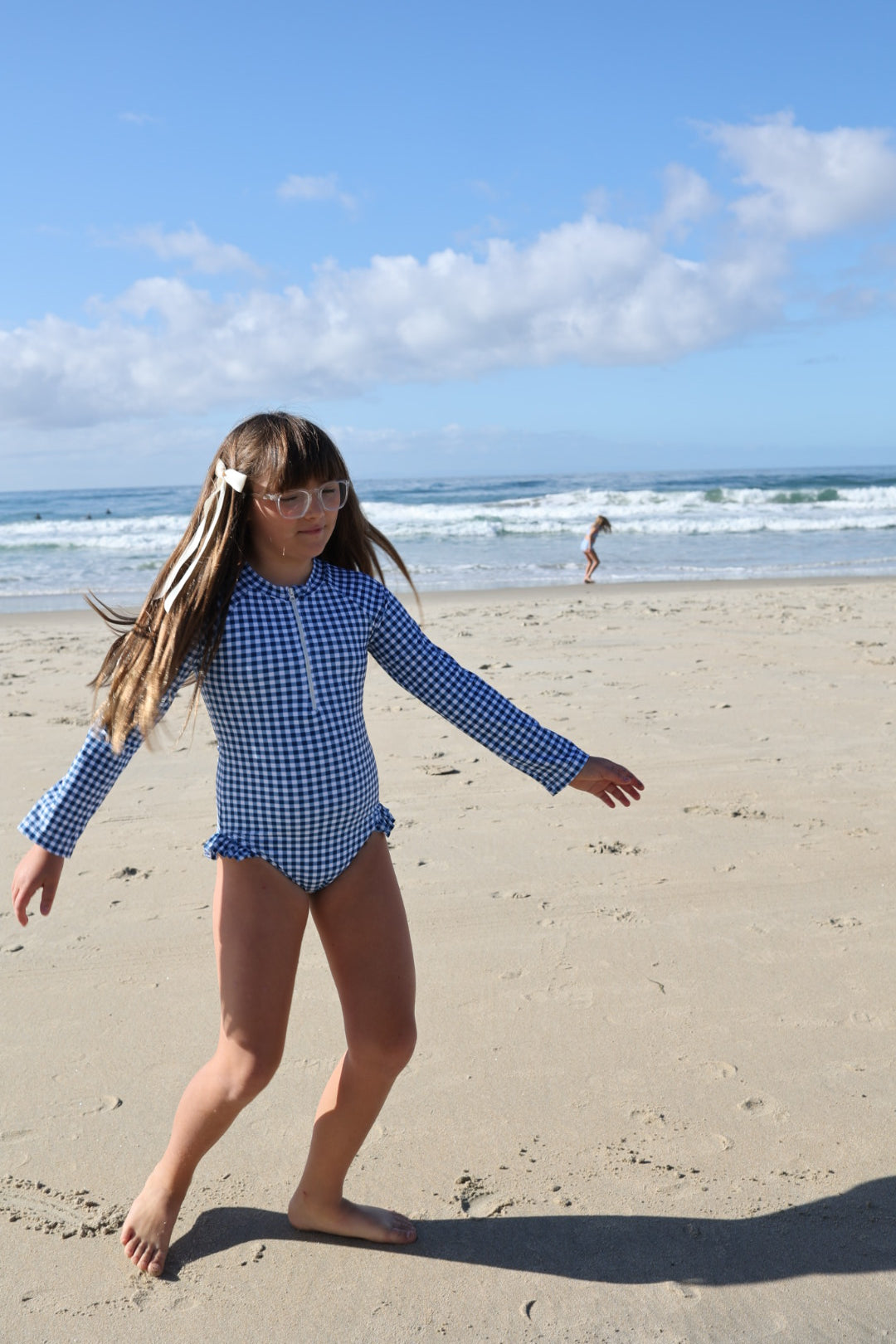 A girl wears the forever french baby Girls Long Sleeve Swimsuit in Blue Gingham, walking barefoot on the sandy beach with waves and a partly cloudy sky. Nearby, another person enjoys summer swimwear at the shoreline.