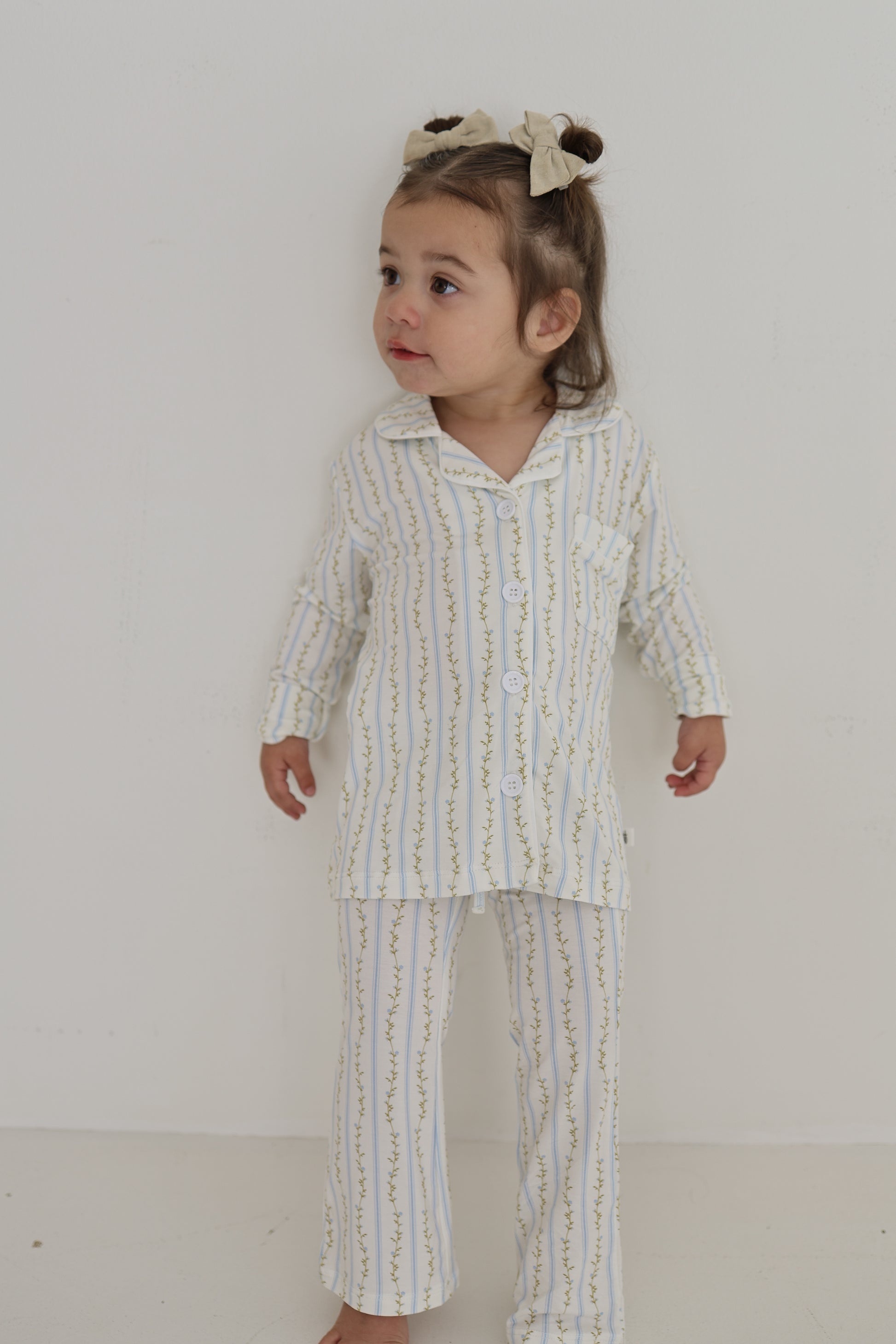 A young child with light brown hair tied in a bow stands barefoot against a white background, wearing the forever french baby Girl’s Bamboo Button Up Lounge Set | Dainty Vines—ultra-soft, hypo-allergenic bamboo clothing with delicate yellow and green vertical stripes.