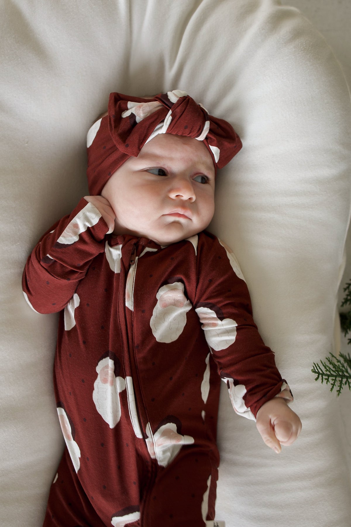 Baby wearing a dark red, long-sleeve Bamboo Zip Pajamas | Vintage Santa sleeper with a matching headband, lying on a white cushion.