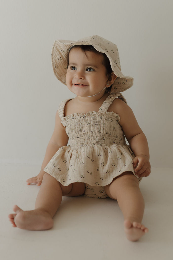 A smiling baby in the forever french baby Sleeveless Muslin Romper & Hat | Flora, featuring light beige fabric and ruffle details, sits on the floor against a plain light background.