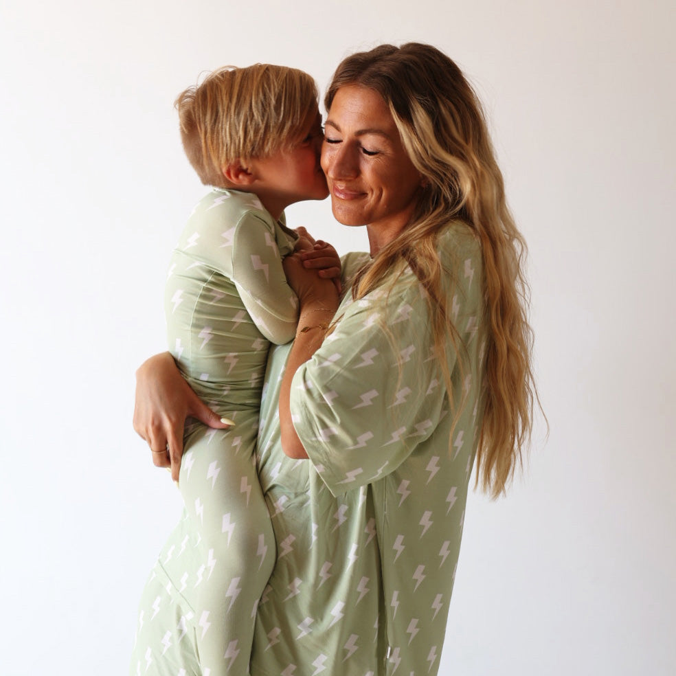 A woman with long, wavy hair holds a child who kisses her cheek. Both wear matching "Flash" light green bamboo short sets by forever french baby, featuring a white lightning bolt pattern against a plain white background.