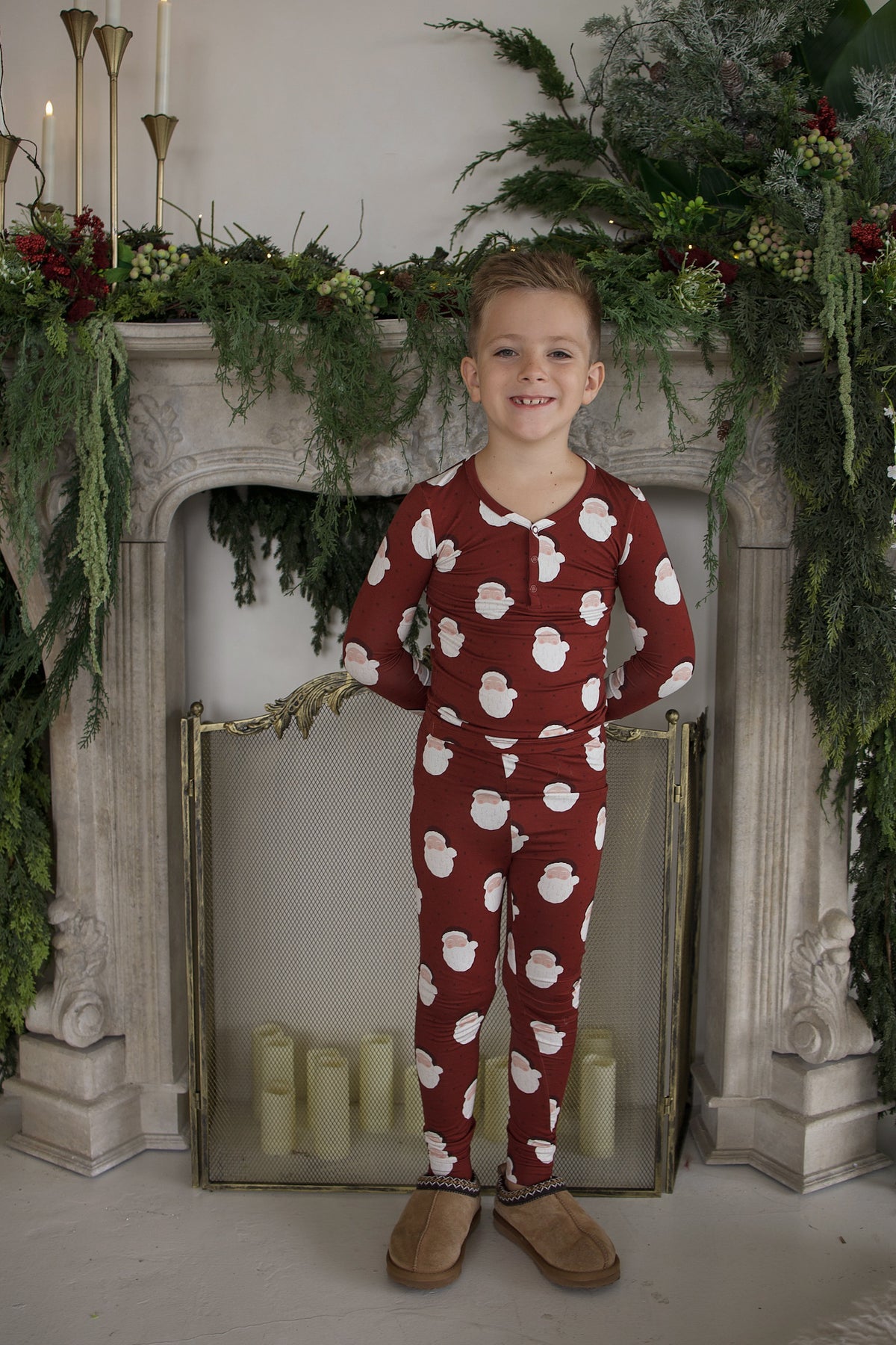 A young boy wearing Bamboo Two Piece Pajamas | Vintage Santa with a white Santa Claus print standing in front of a fireplace decorated with greenery.