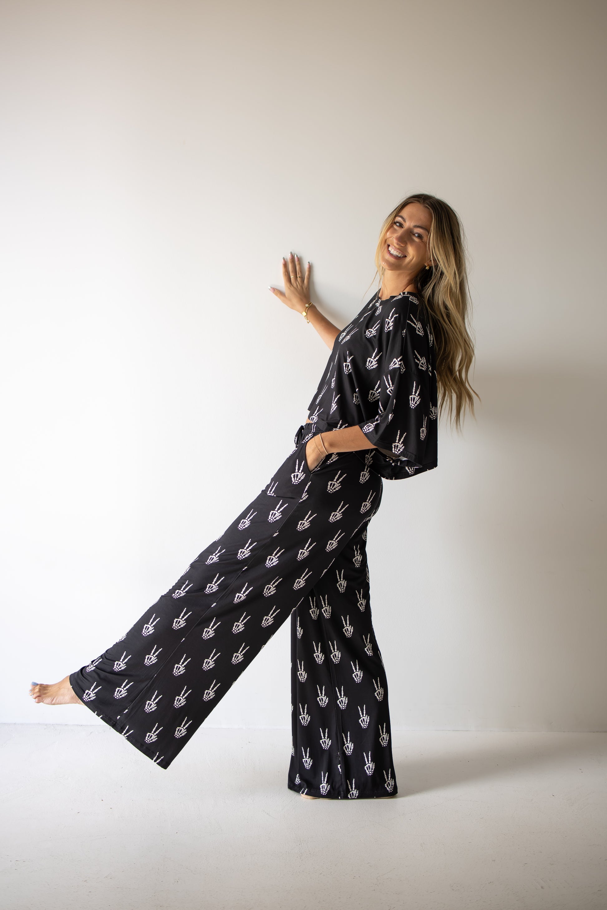 A smiling woman stands barefoot against a plain wall, one foot raised, wearing the forever french baby Women’s Bamboo Crop Tee Lounge Set in Midnight Skelly—with white handprint designs—her left hand on the wall and her right hand in her pocket.
