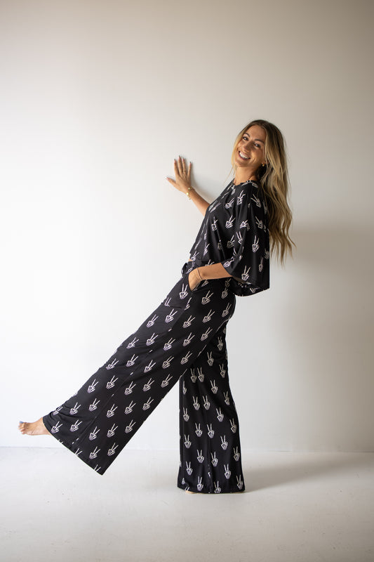 A smiling woman stands barefoot against a plain wall, one foot raised, wearing the forever french baby Women’s Bamboo Crop Tee Lounge Set in Midnight Skelly—with white handprint designs—her left hand on the wall and her right hand in her pocket.