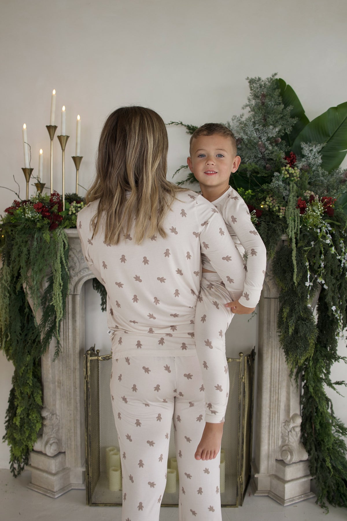 A woman and young boy in matching Bamboo Two Piece Pajamas | Gingersnap with a brown gingerbread man pattern, posed in front of a fireplace decorated with greenery and candles.