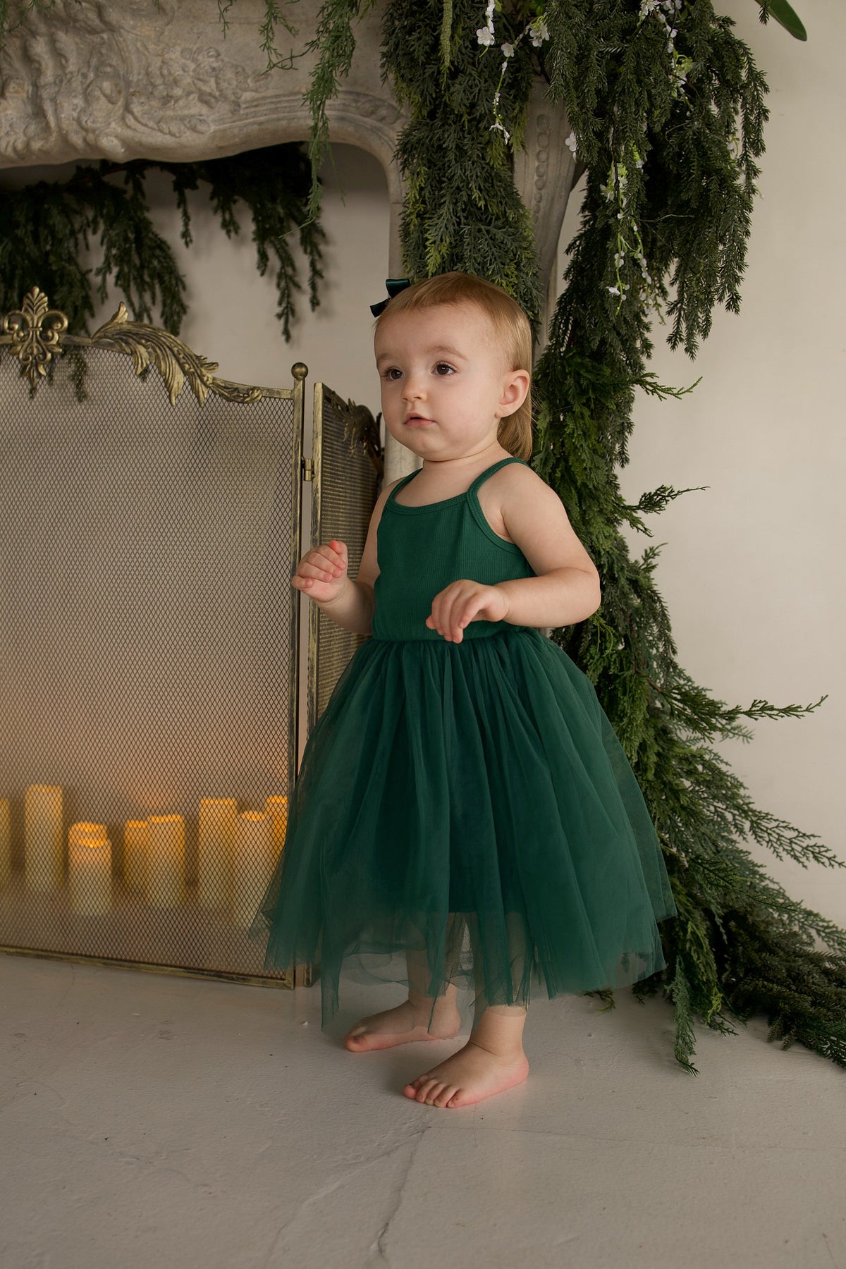 A toddler stands in a dark green forever french baby ff Tutu Dress | Mistletoe with a layered tulle skirt in front of a fireplace with lit candles.