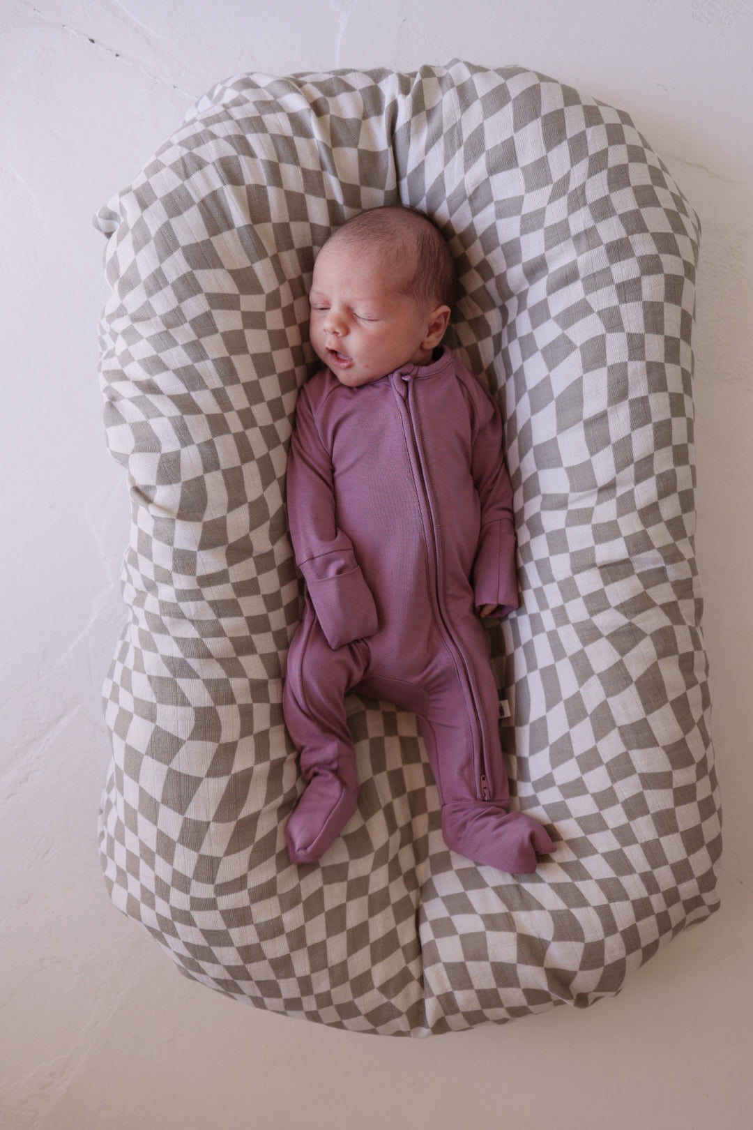 An infant in hypo-allergenic Bamboo Zip Pajamas | Amethyst from forever french baby sleeps peacefully on a cushioned checkered baby lounger. The lounger features a pattern of alternating light and dark squares, creating a soft, cozy environment, with the background being a plain, light-colored floor.