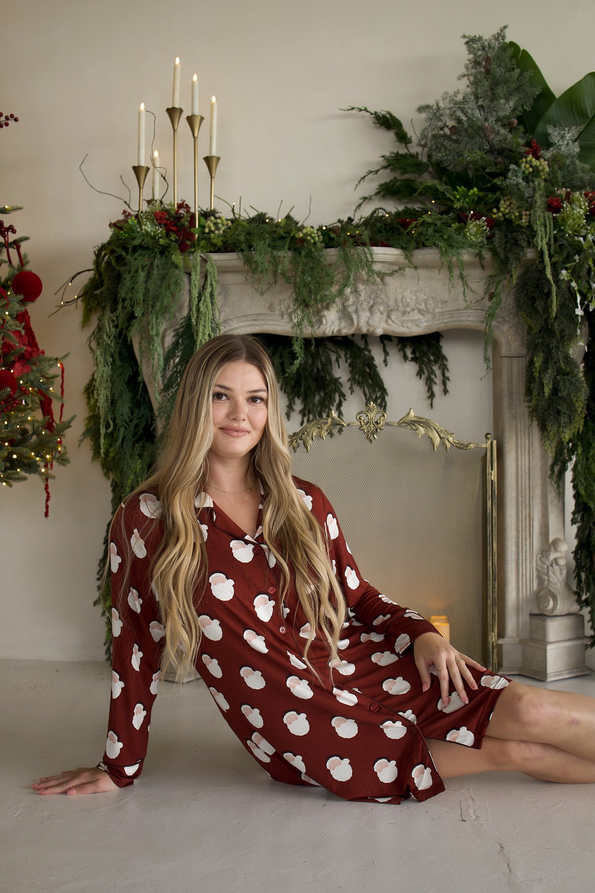 A woman sits on the floor wearing a forever french baby Women’s Bamboo Sleeping Dress | Vintage Santa, set against a fireplace decorated for Christmas.