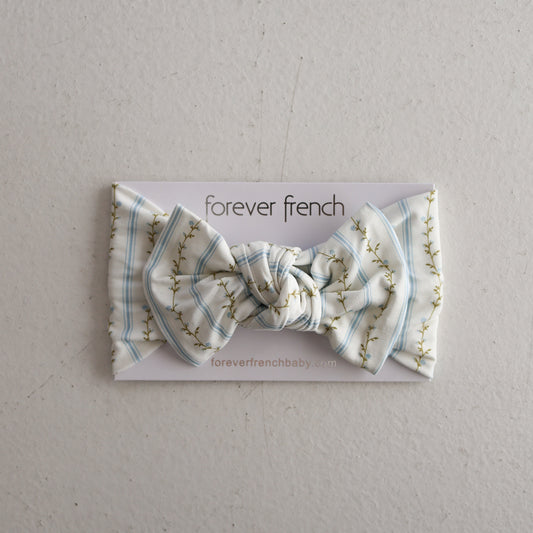 The Bamboo Head Wrap | Dainty Vines by forever french baby features a light blue and white plaid design with a delicate floral pattern, displayed on branded packaging against a light gray textured background.