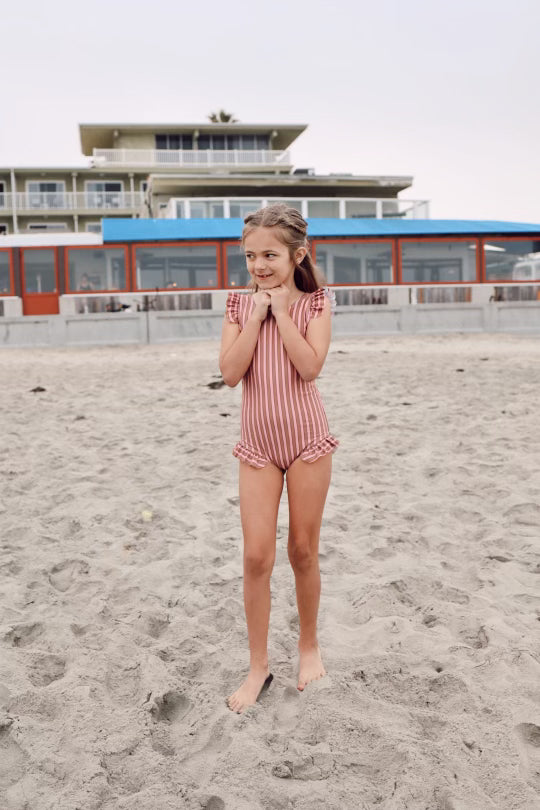 A young girl stands barefoot on a sandy beach, smiling with her arms held close to her chest, wearing the forever french baby One Piece Girls Swim Suit | Boardwalk in pink and white stripes. Behind her, modern beachfront buildings with large windows and balconies can be seen. Her swimsuit provides UPF 50+ protection against the overcast sky.