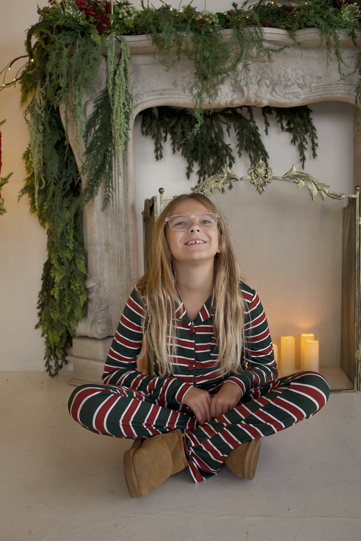A girl sits cross-legged on the floor in front of a fireplace wearing a Girl’s Bamboo Button Up Lounge Set | Holiday Stripe.