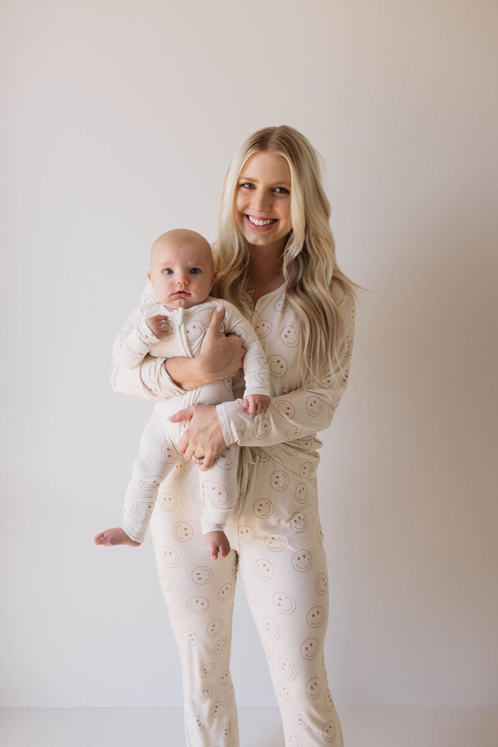 A woman with long blonde hair stands against a plain backdrop, smiling and holding a baby. Both are dressed in matching Women's Bamboo Pajamas from forever french baby, featuring the Just Smile pattern. The woman, ideal for breastfeeding moms, is looking at the camera while the baby rests comfortably in her arms wearing cozy pajamas made of bamboo viscose.