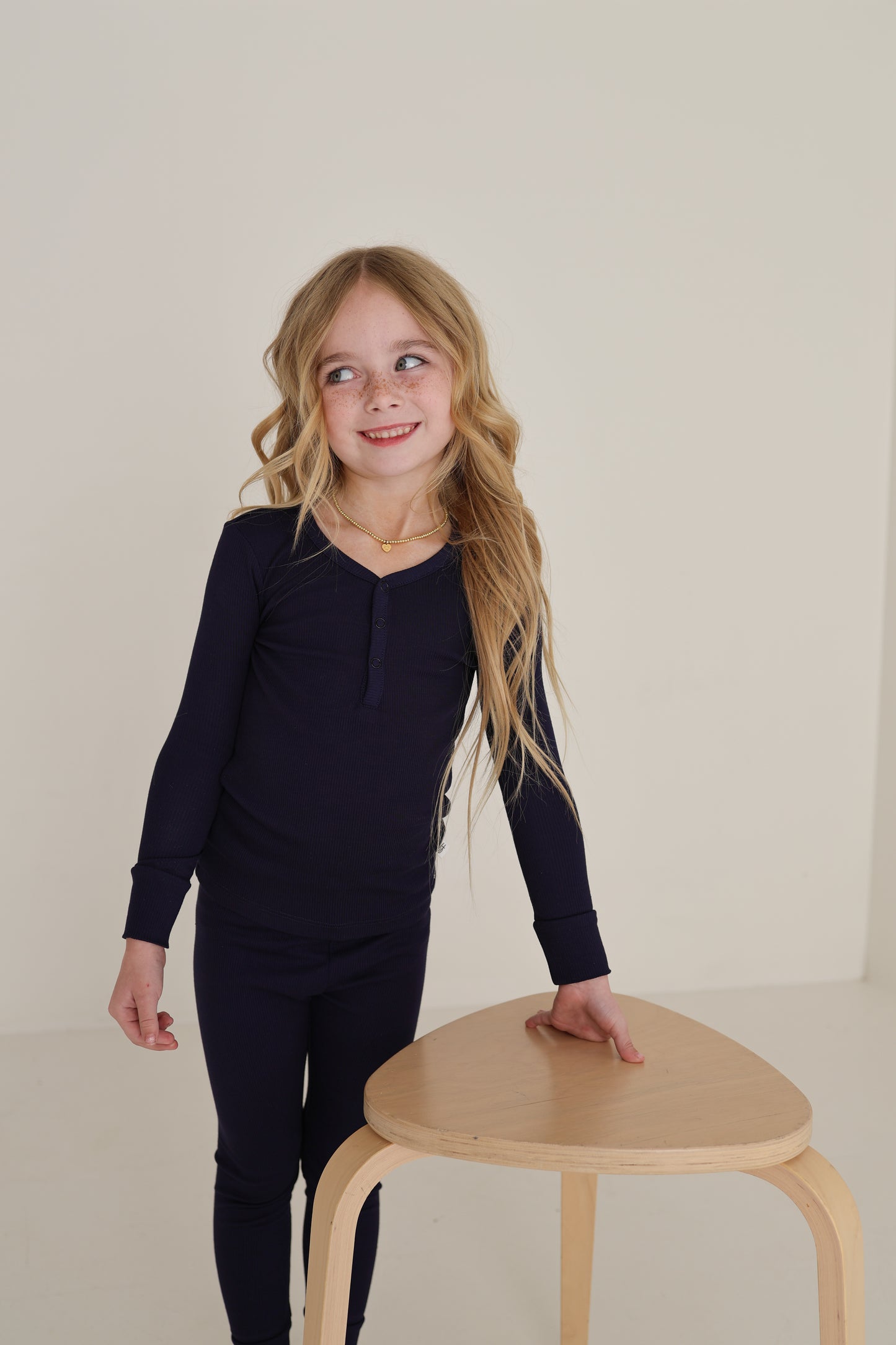 A young girl with long blonde hair, wearing forever french baby Bamboo Two Piece Pajamas in Blueberry Ribbed, stands by a light wooden stool and smiles to the side against a plain light background.