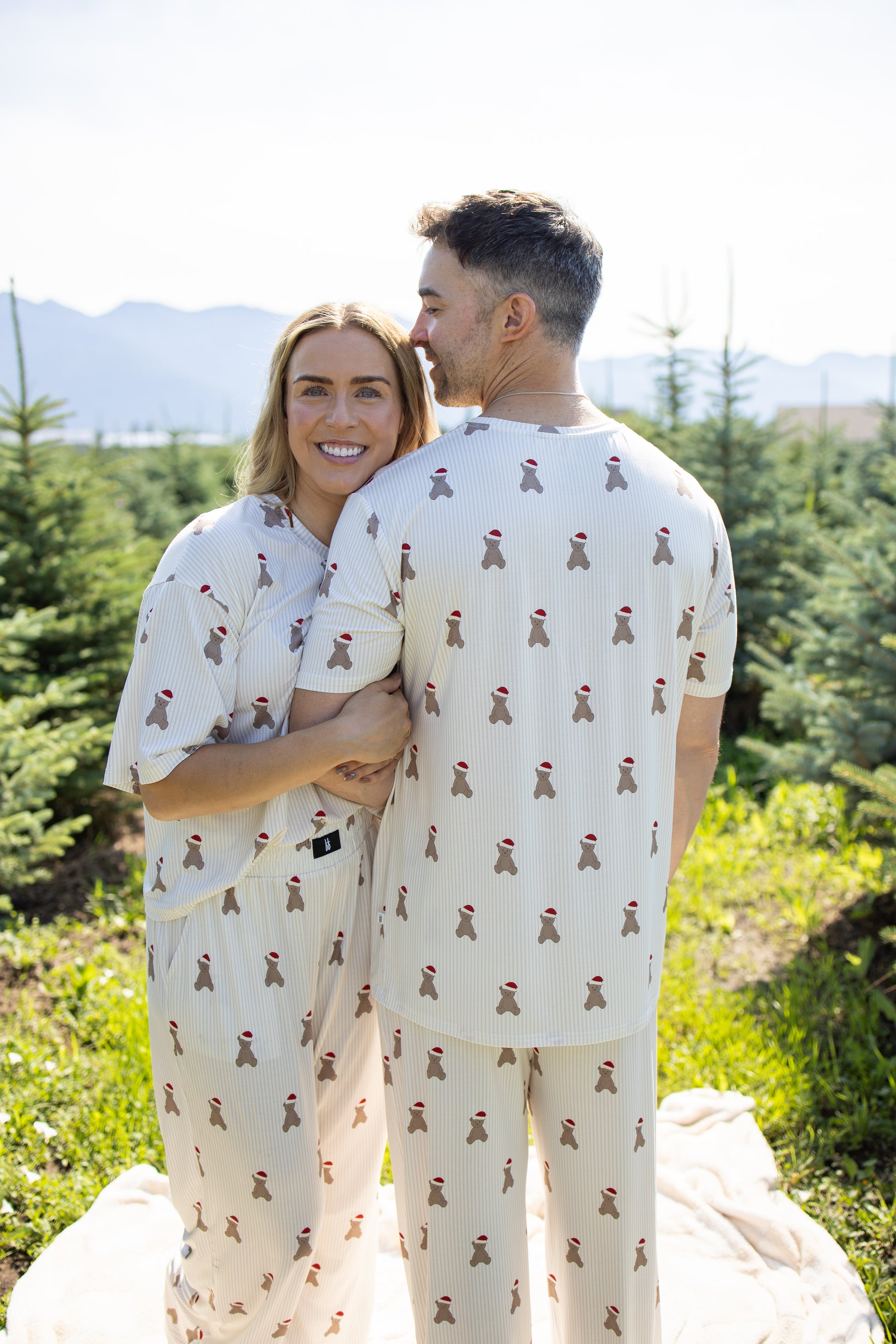 A smiling couple stands close together outdoors among pine trees on a sunny day, enjoying the comfort of forever french baby's Men's Short Sleeve Pajamas | Santa Teddy—hypoallergenic sleepwear crafted from breathable bamboo, featuring a matching cream design with small red figures.
