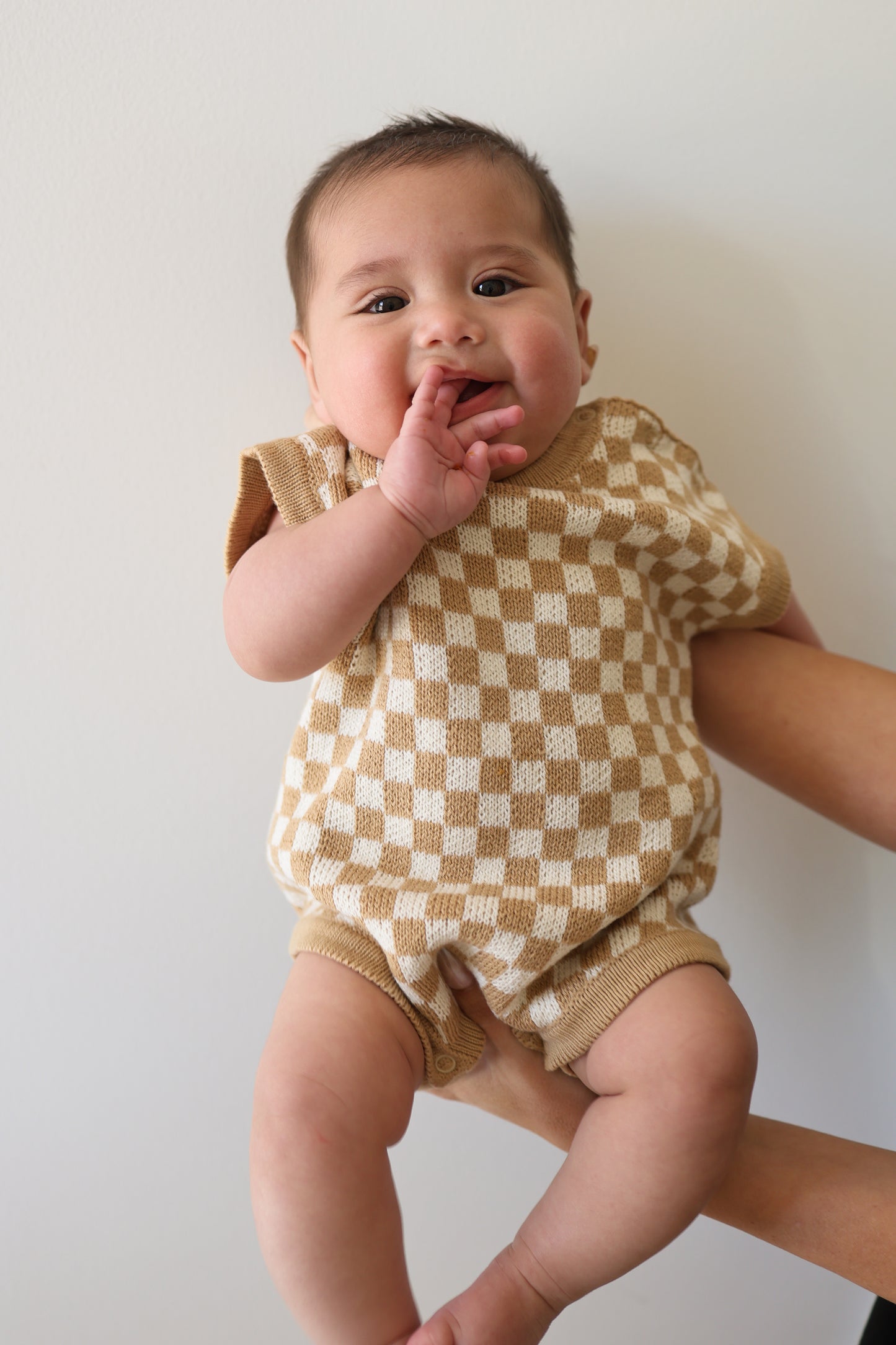 A baby in the forever french baby Knit Romper | Latte Check is held up against a plain light background, looking at the camera with one hand near their mouth.