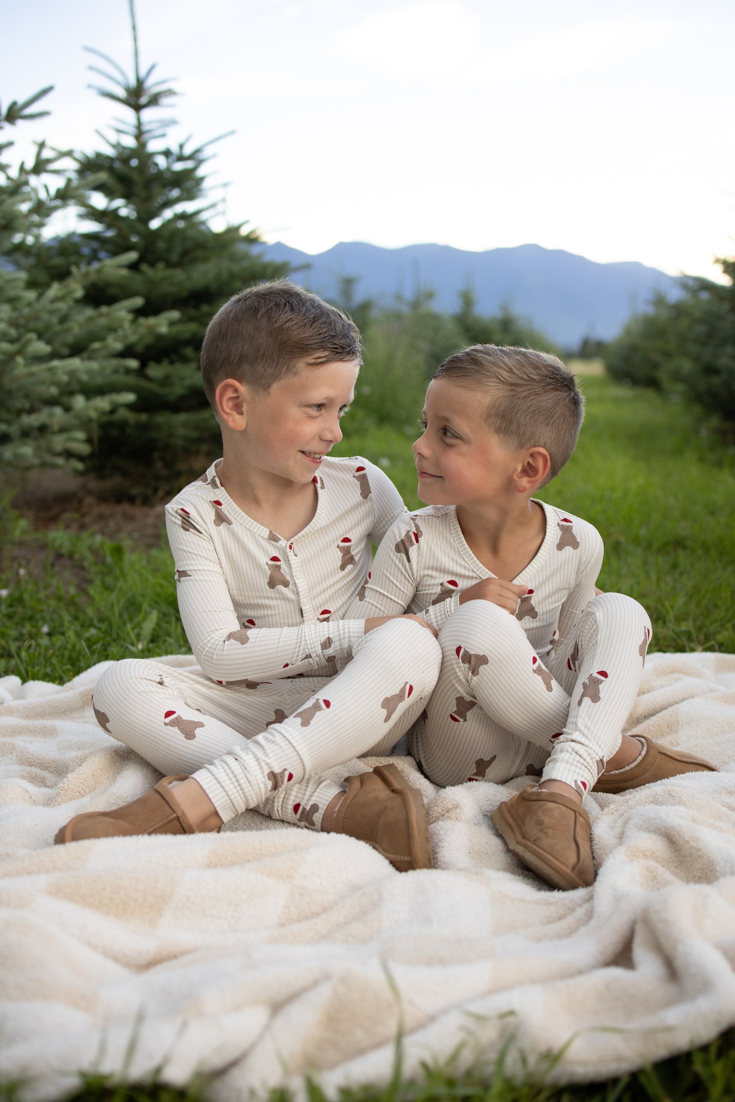 Two young boys in matching forever french baby Bamboo Two Piece Pajamas | Santa Teddy sit together on a blanket outdoors, smiling at each other with pine trees and mountains in the background.