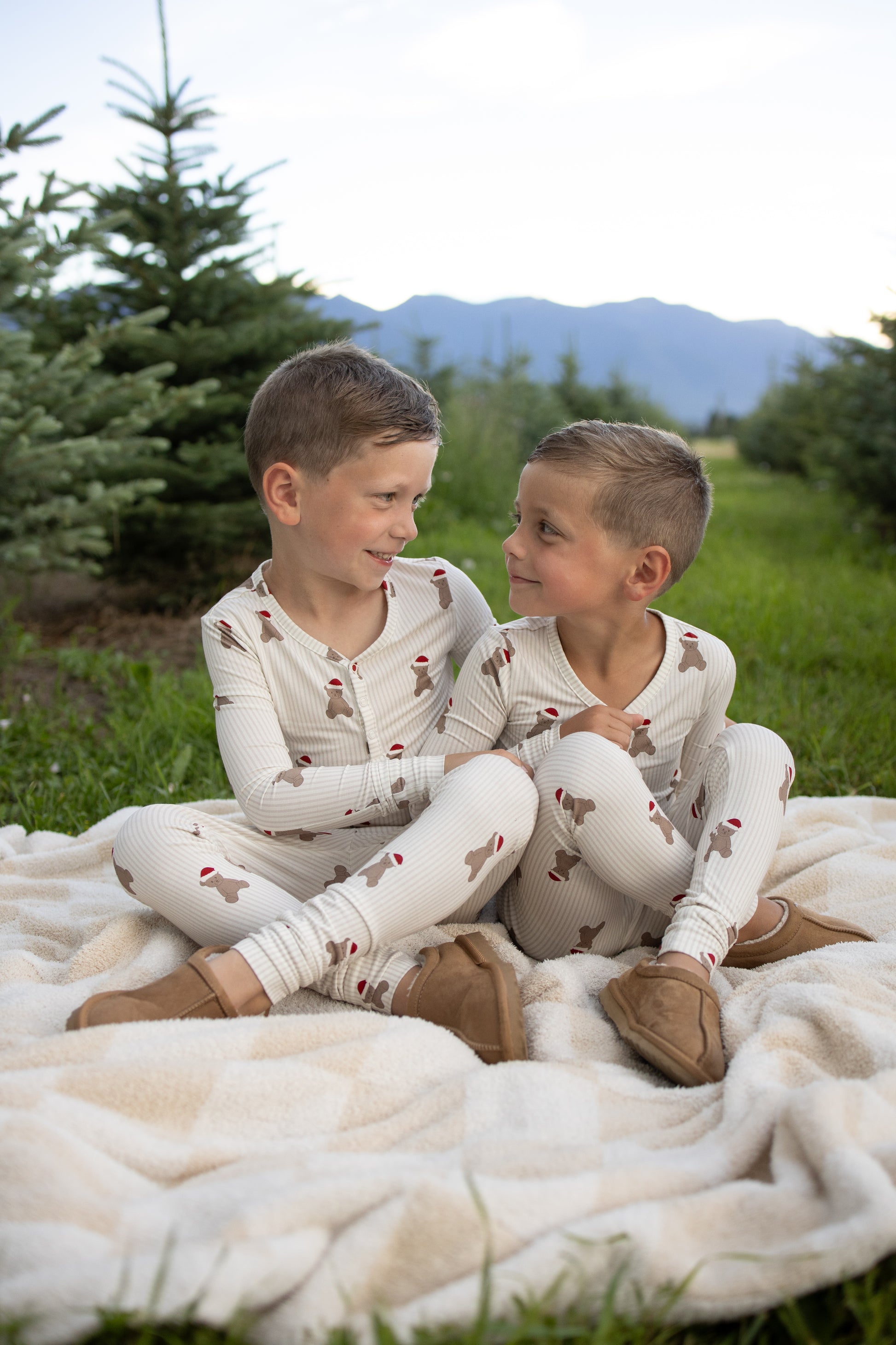 Two young boys in matching forever french baby Bamboo Two Piece Pajamas | Santa Teddy sit together on a blanket outdoors, smiling at each other with pine trees and mountains in the background.
