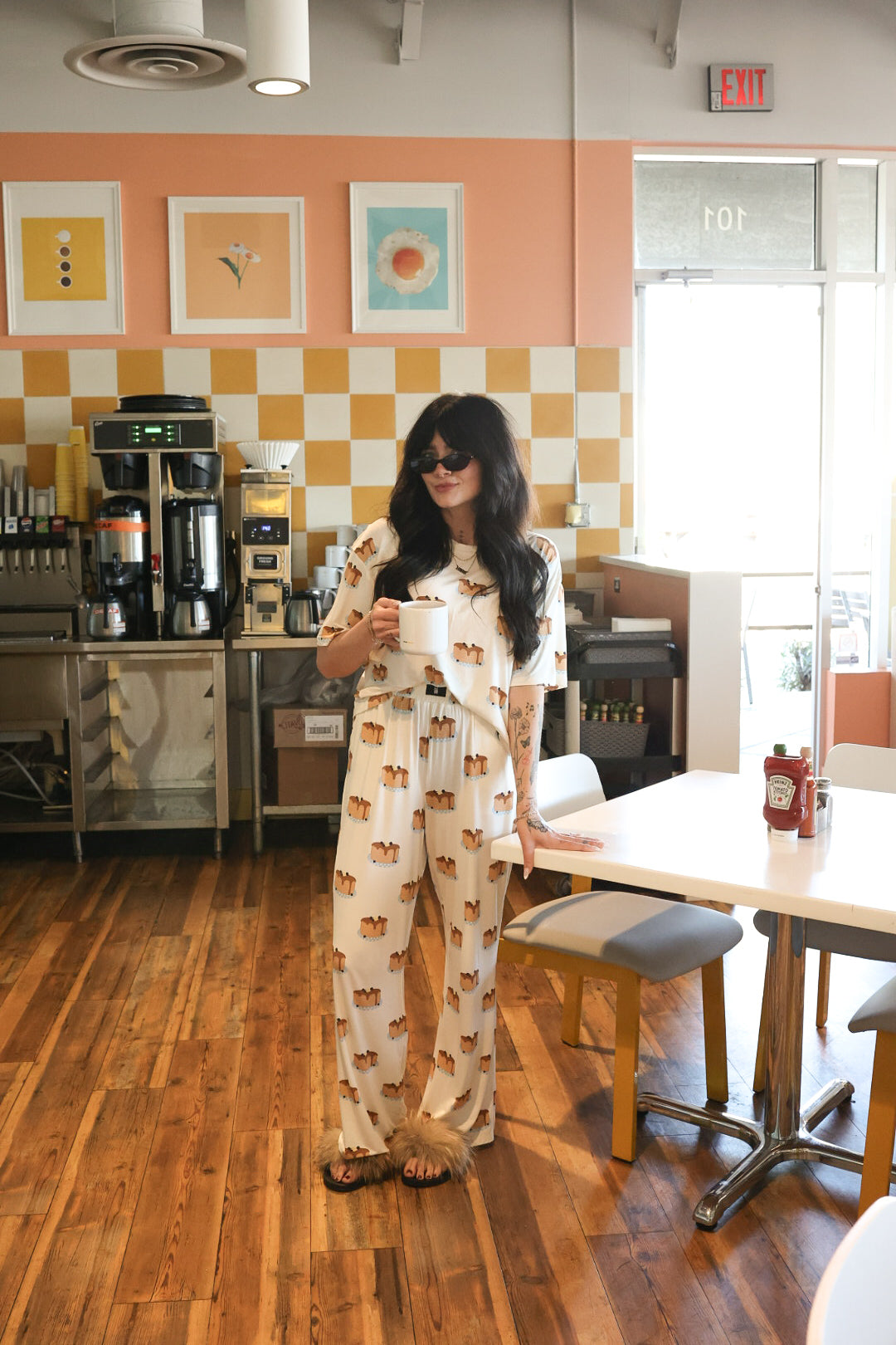 A woman with long dark hair in sunglasses wears the Women's Boxy Tee Lounge Set by forever french baby, standing in a bright retro café with wood floors, yellow-checkered walls, and framed art, holding a white mug.