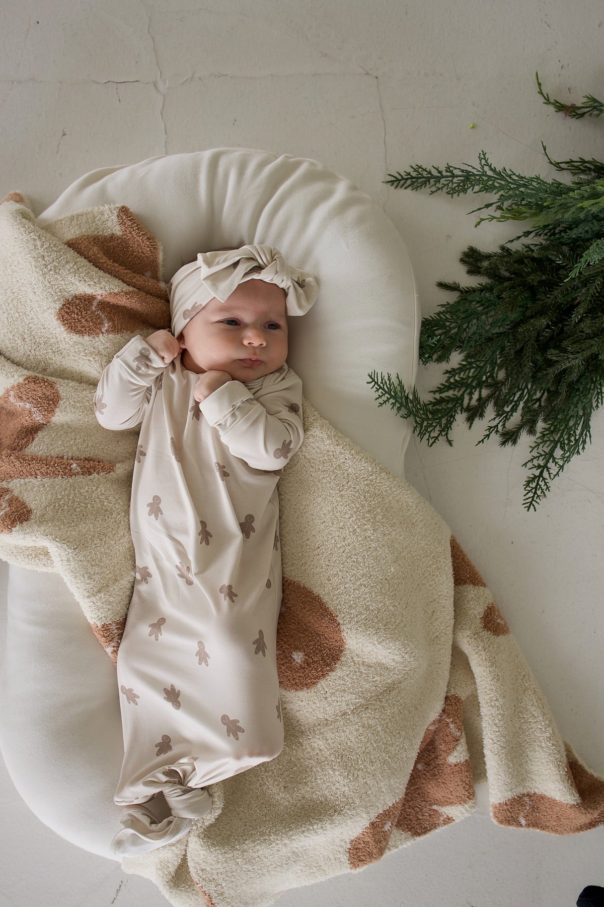 A baby wearing a beige Bamboo Knotted Gown | Gingersnap and matching headband, lying in a lounger on a patterned blanket next to evergreen branches.