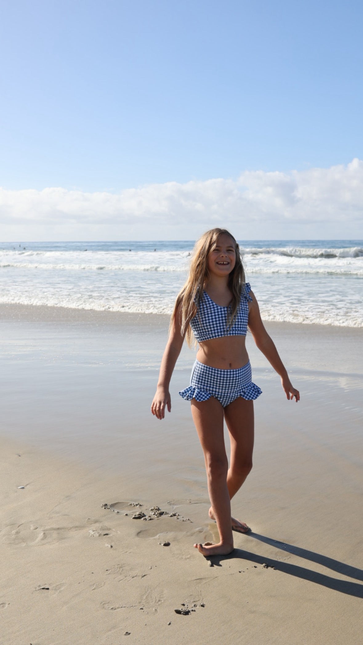 A young girl smiles and walks on a sandy beach in the forever french baby Girls Two Piece Swimsuit | Blue Gingham, with gentle waves and a partly cloudy sky in the background.