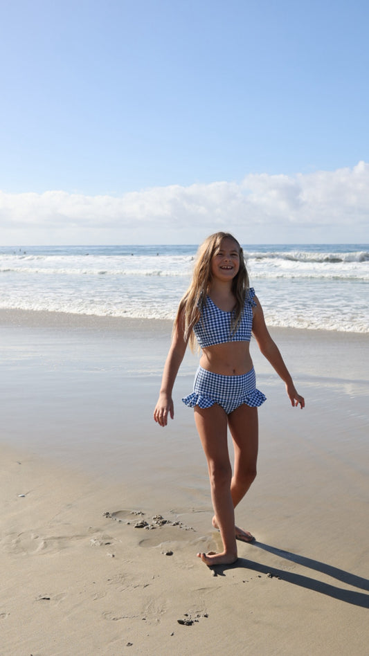 A young girl smiles and walks on a sandy beach in the forever french baby Girls Two Piece Swimsuit | Blue Gingham, with gentle waves and a partly cloudy sky in the background.