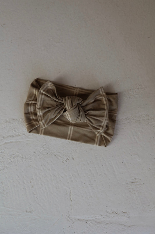A beige Bamboo Head Wrap | Linen Grid with a large knotted bow from forever french baby, featuring a plaid pattern, is displayed on a textured off-white surface.