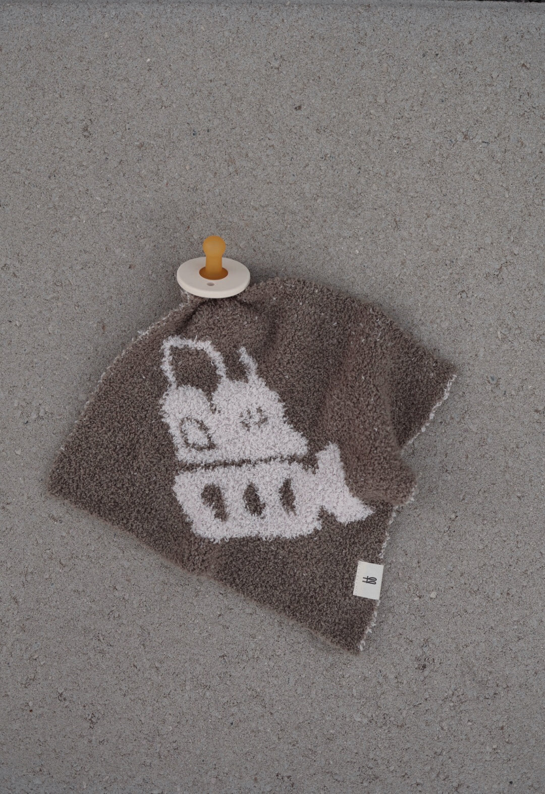 A brown "Lovey | Bulldozer" by forever french baby featuring a white cartoon bulldozer design rests on a gray surface, with a beige pacifier with brown nipple attached to one corner.