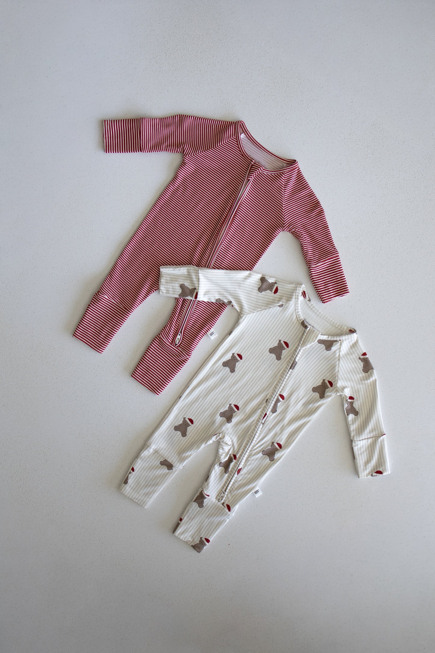 Two forever french baby Bamboo Zip Pajamas | Santa Teddy are displayed on a light background: one features a red base with thin white stripes, while the other is white with subtle red accents and an adorable Santa Teddy print. Both pajamas have long sleeves and legs, front zippers for easy dressing, and are crafted from hypoallergenic material to suit delicate baby skin.
