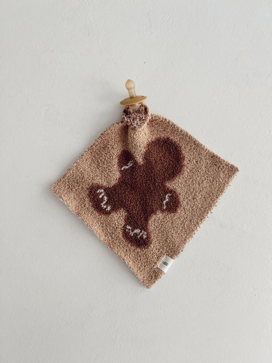 A plush Lovey from the Gingerbread Collection by forever french baby, featuring a delightful gingerbread man design, is crafted from soft microfiber feather yarn and is attached to a beige pacifier, set against a white background.