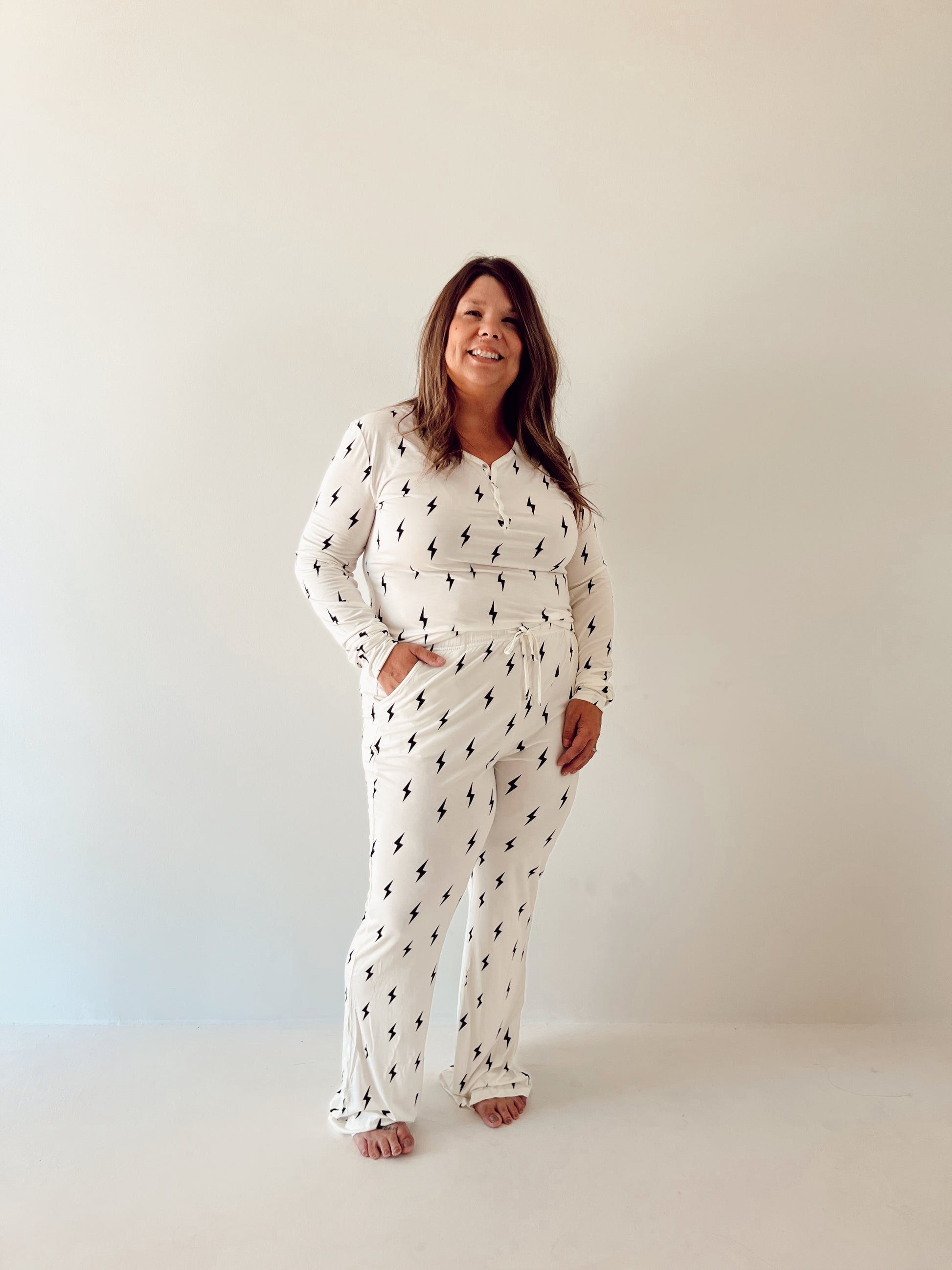 A woman with long, wavy hair is standing barefoot in front of a plain, light-colored wall. She is smiling and wearing forever french baby's Women's Bamboo Pajamas in a White & Black Lightning Bolt pattern. Her hands are in her pockets.