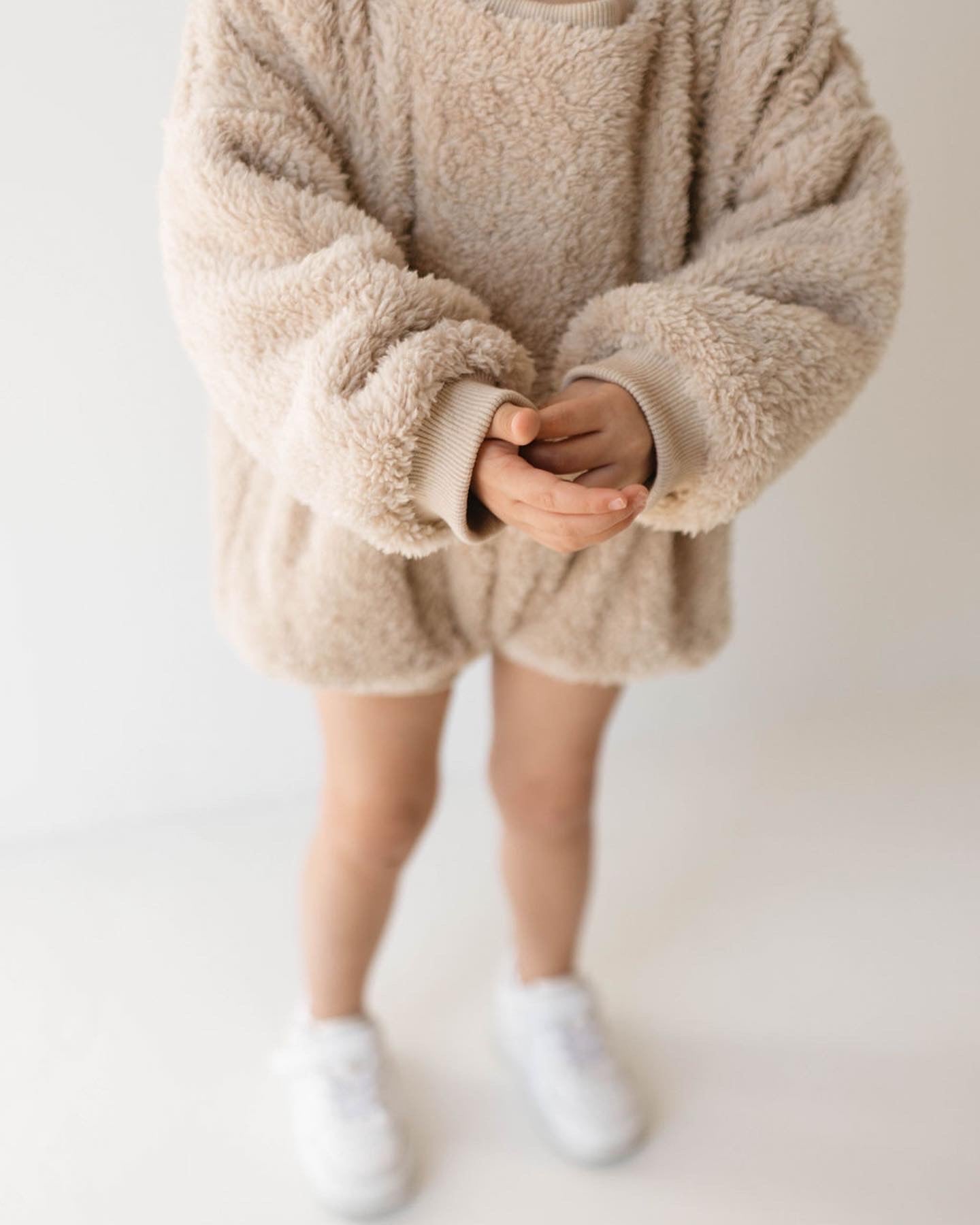 A young child wearing an oversized, fluffy beige sweater stands against a plain background. Clasping their hands together at their chest, they are dressed in a cozy romper from forever french baby, which looks as comfortable as the Long Sleeve Romper | Teddy Bear. White sneakers peek out from below the sweater.