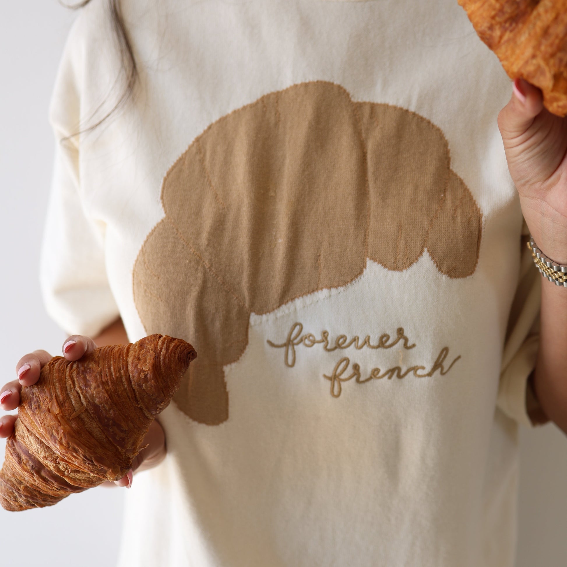 A person wears the forever french baby Women's Knit Pant Set | Croissants, featuring a croissant graphic and “forever french” text, while holding croissants in each hand. The background is simple and light.