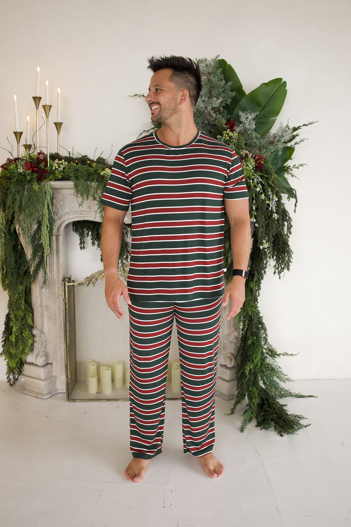 A man wears a hypoallergenic Men’s Short Sleeve Pajamas | Holiday Stripe set, featuring a short-sleeve top and pants in a red, white, and green horizontal stripe pattern.