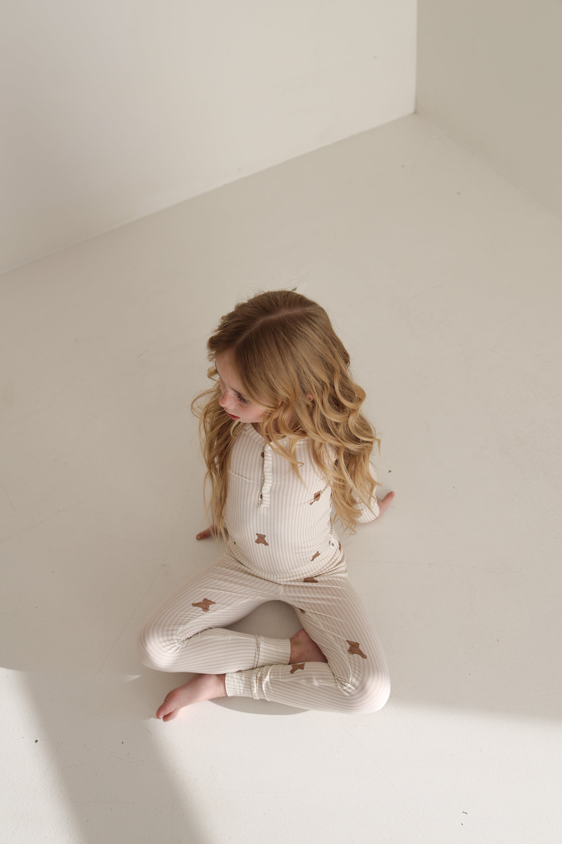 A young child with long, wavy hair sits barefoot on a white floor in forever french baby’s Bamboo Two Piece Pajamas | Teddy, featuring a light-colored star pattern, gazing off to the side as sunlight brightens the nearby floor.