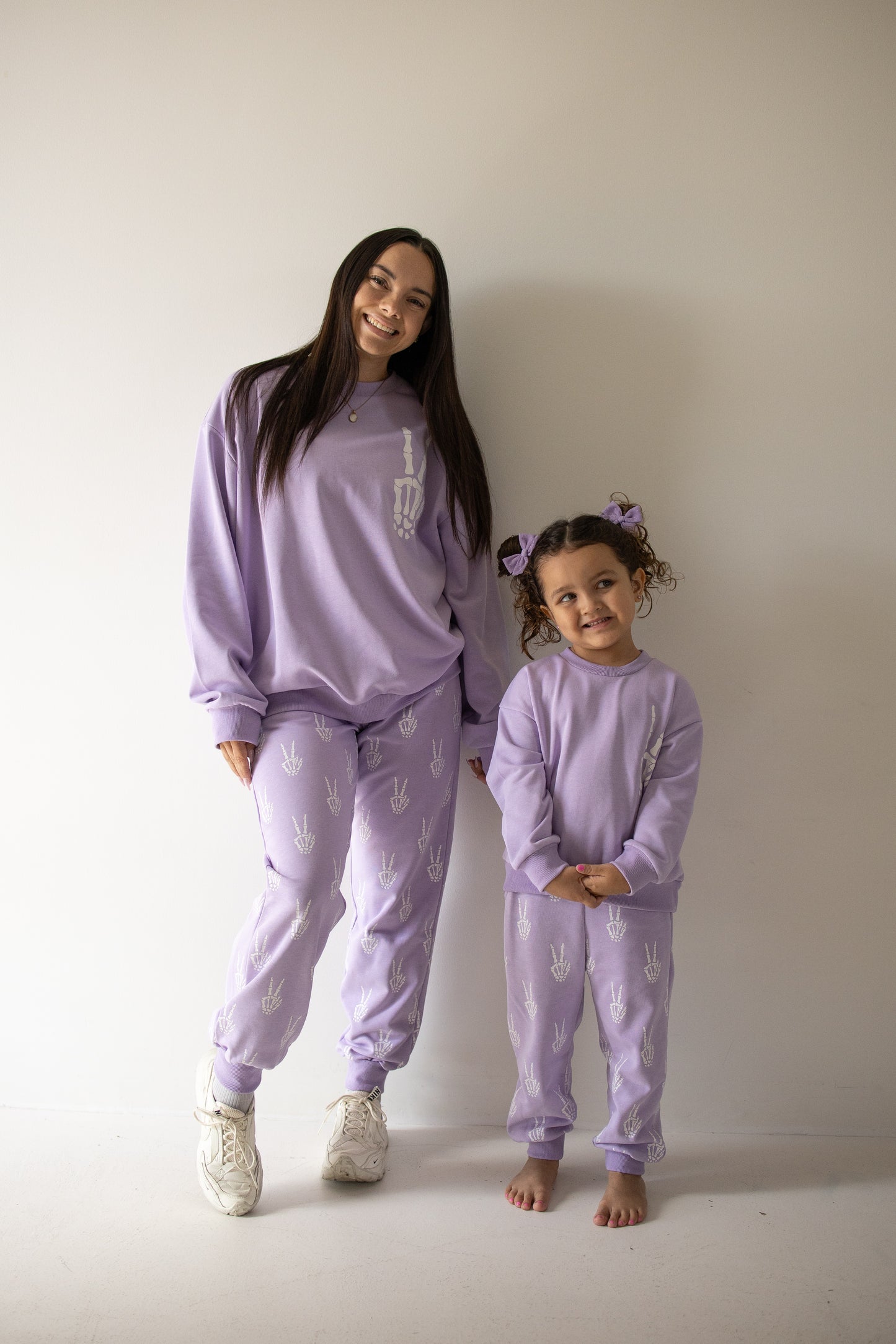 An adult and a child stand side by side wearing matching forever french baby Women’s Sweat Set | Enchanted Skelly sweatsuits in lavender, featuring white Enchanted Skelly designs. The adult smiles warmly while the barefoot child, adorned with hair bows, faces forward beside her. Together they perfectly capture the whimsy and charm of spooky season style against a simple light background.