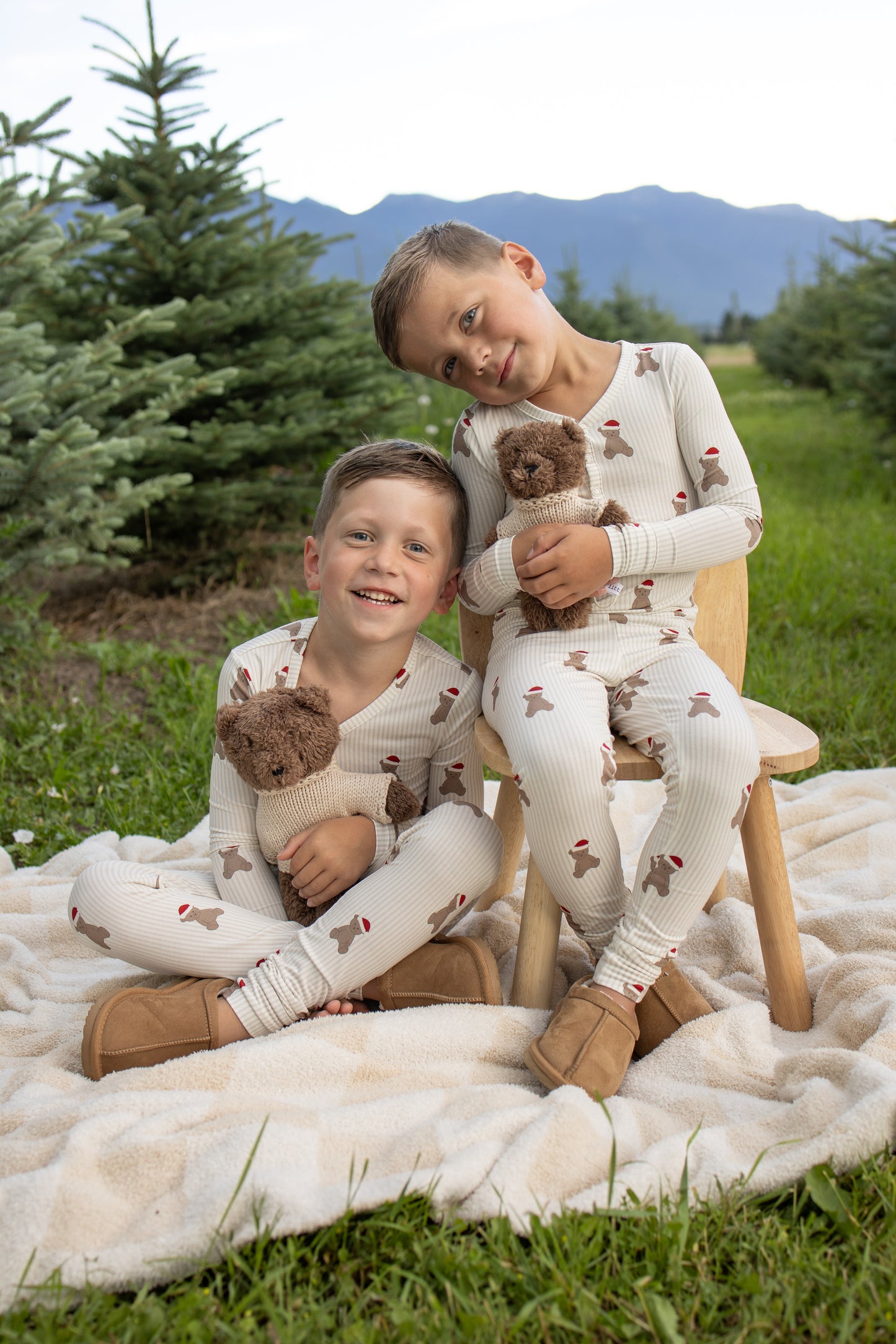 Two young boys sit outdoors on a blanket among evergreen trees and mountains, each holding teddy bears. Both wear matching forever french baby Bamboo Two Piece Pajamas in the charming Santa Teddy print, paired with boots. One boy sits on a small wooden chair, leaning his head against the other as they enjoy cozy moments in their hypoallergenic kids sleepwear.