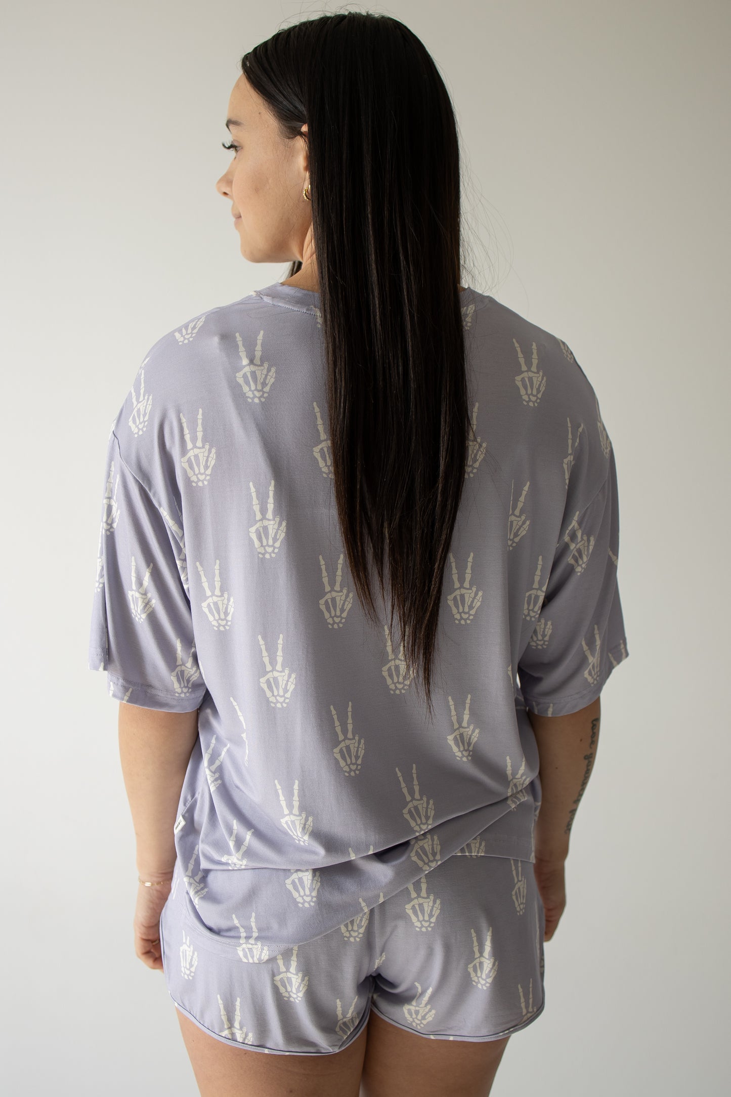 A woman with long dark hair faces away, wearing the forever french baby Women’s Bamboo Short Set in the Enchanted Skelly print—a matching light purple pajama set adorned with a white hand peace sign pattern—standing against a plain light background.