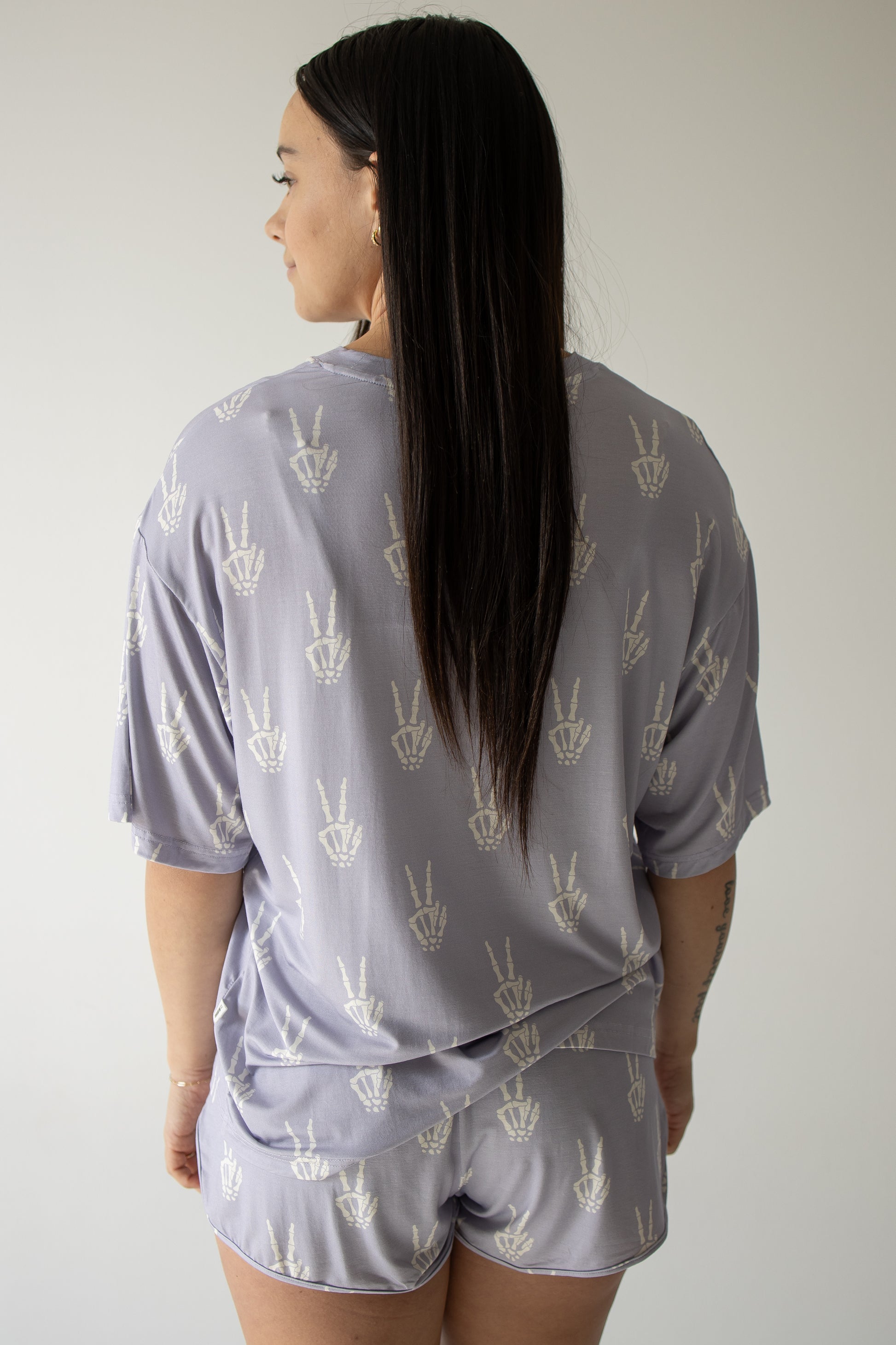 A woman with long dark hair faces away, wearing the forever french baby Women’s Bamboo Short Set in the Enchanted Skelly print—a matching light purple pajama set adorned with a white hand peace sign pattern—standing against a plain light background.