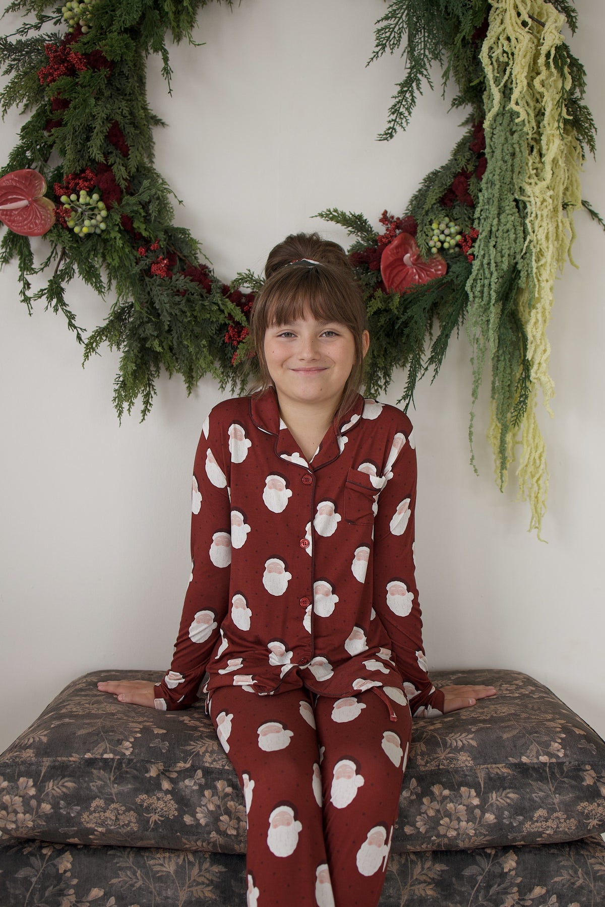 A girl wearing a forever french baby Girl’s Bamboo Button Up Lounge Set in Vintage Santa sits on a patterned ottoman in front of a holiday wreath.