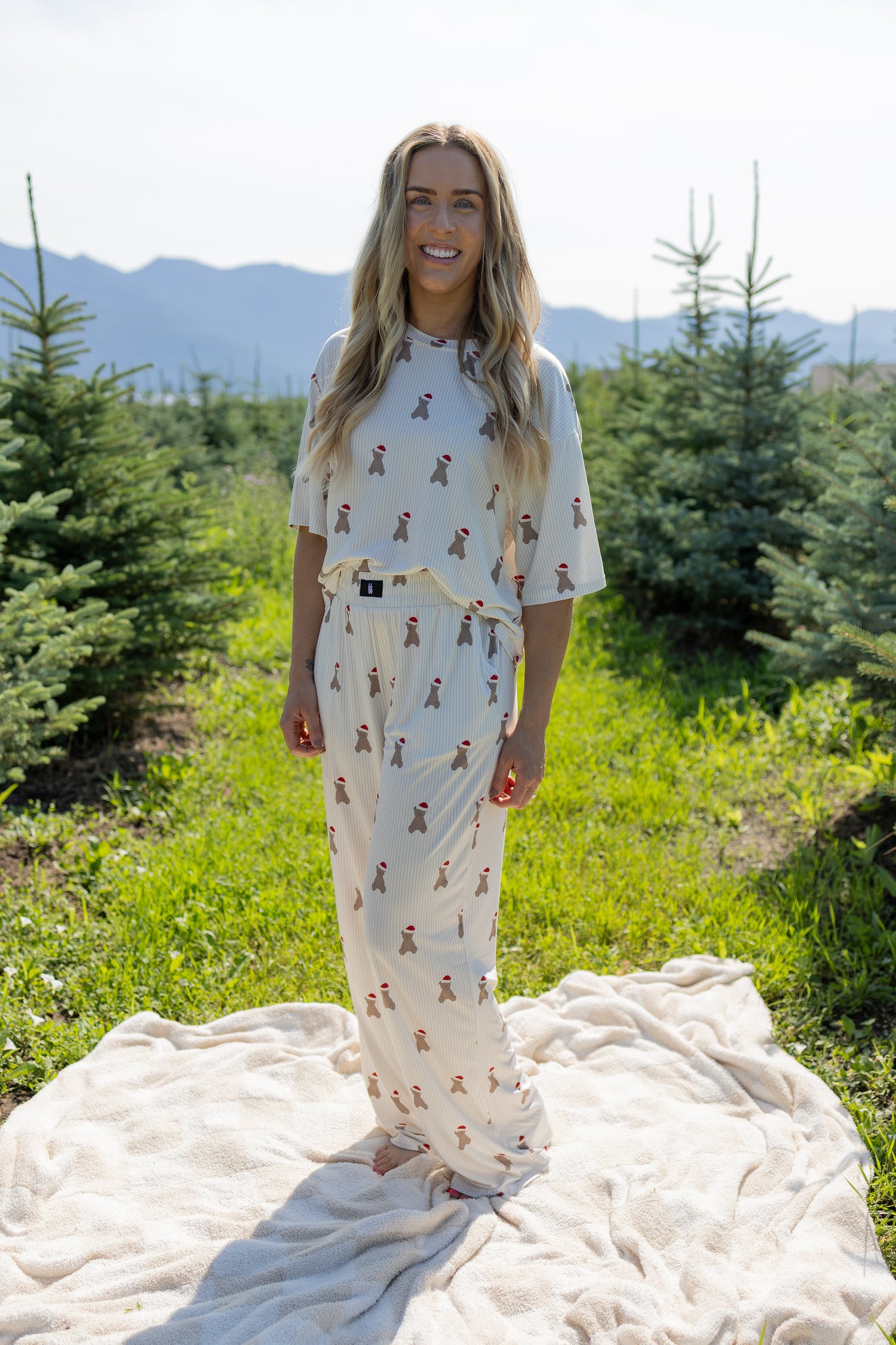 A woman stands smiling on a blanket outdoors among green trees, wearing the Women's Boxy Tee Lounge Set in Santa Teddy by forever french baby—a playful, breathable loungewear ensemble. Mountains are visible in the background under a bright, sunny sky.