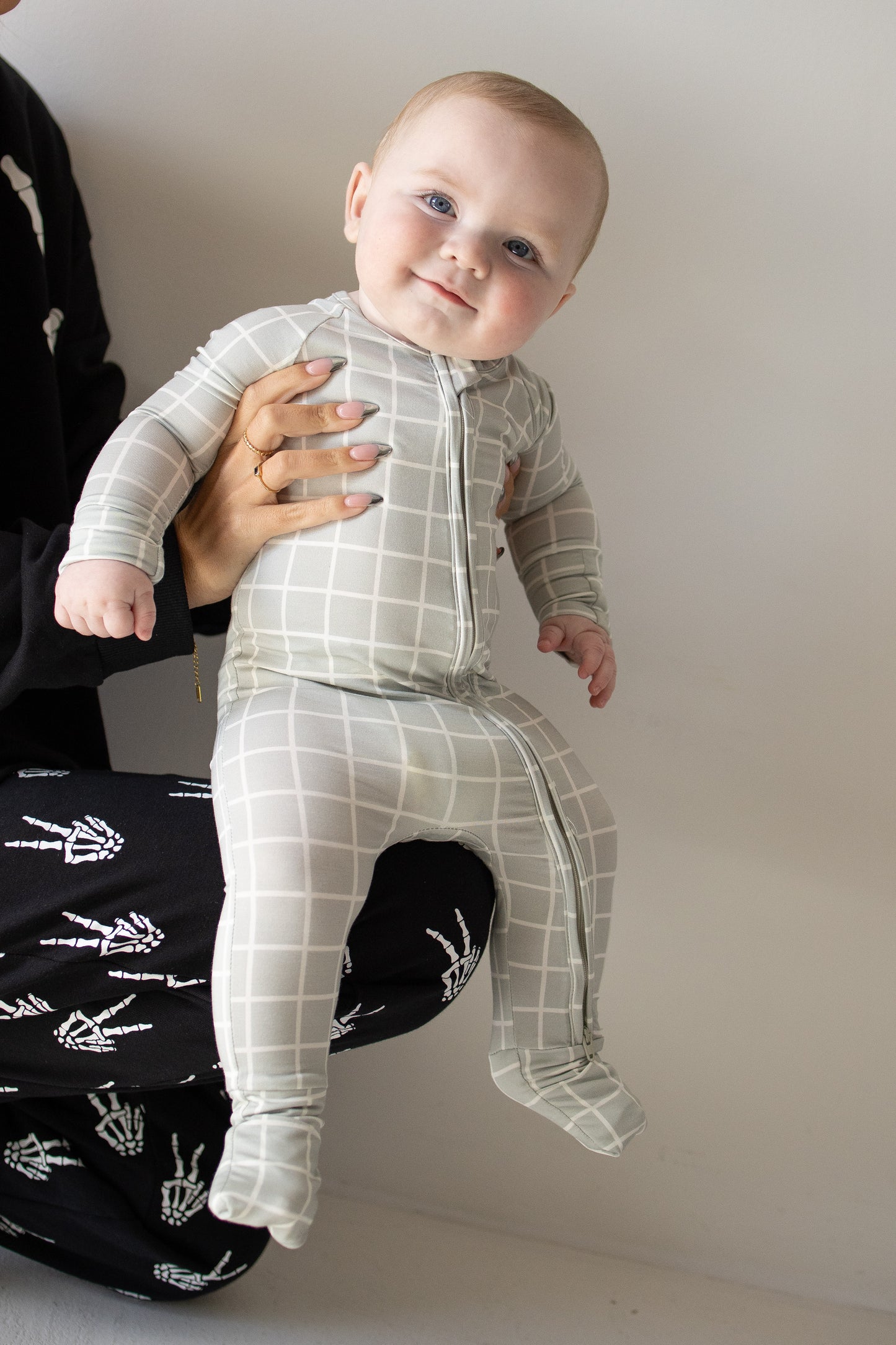 A smiling baby wearing the forever french baby Bamboo Zip Pajamas in Foggy Grid—a light gray, grid-patterned, hypoallergenic onesie—is held up by an adult dressed in black pants with white skeleton hand prints. The baby gazes at the camera with a slight head tilt.