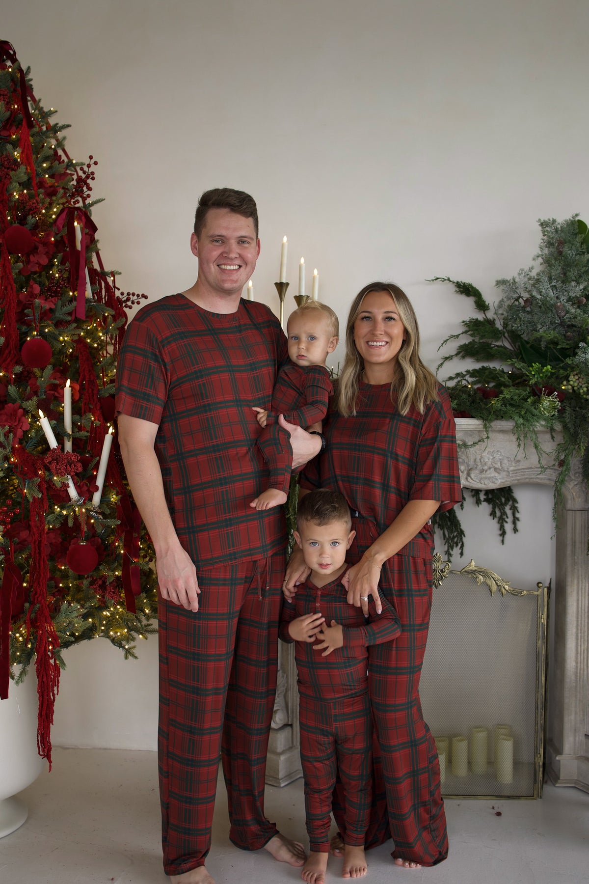 A family of four stands together in front of a Christmas tree and fireplace, all wearing matching Women’s Boxy Tee Lounge Set | Christmas Plaid pajamas.
