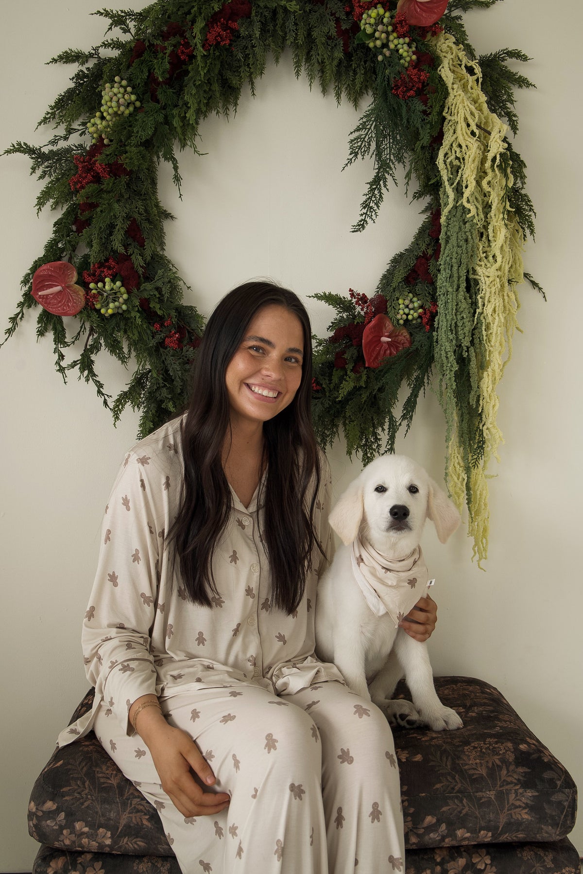 A smiling woman sits next to a white puppy wearing a light-colored Bamboo Bandana | Gingersnap in front of a holiday wreath.
